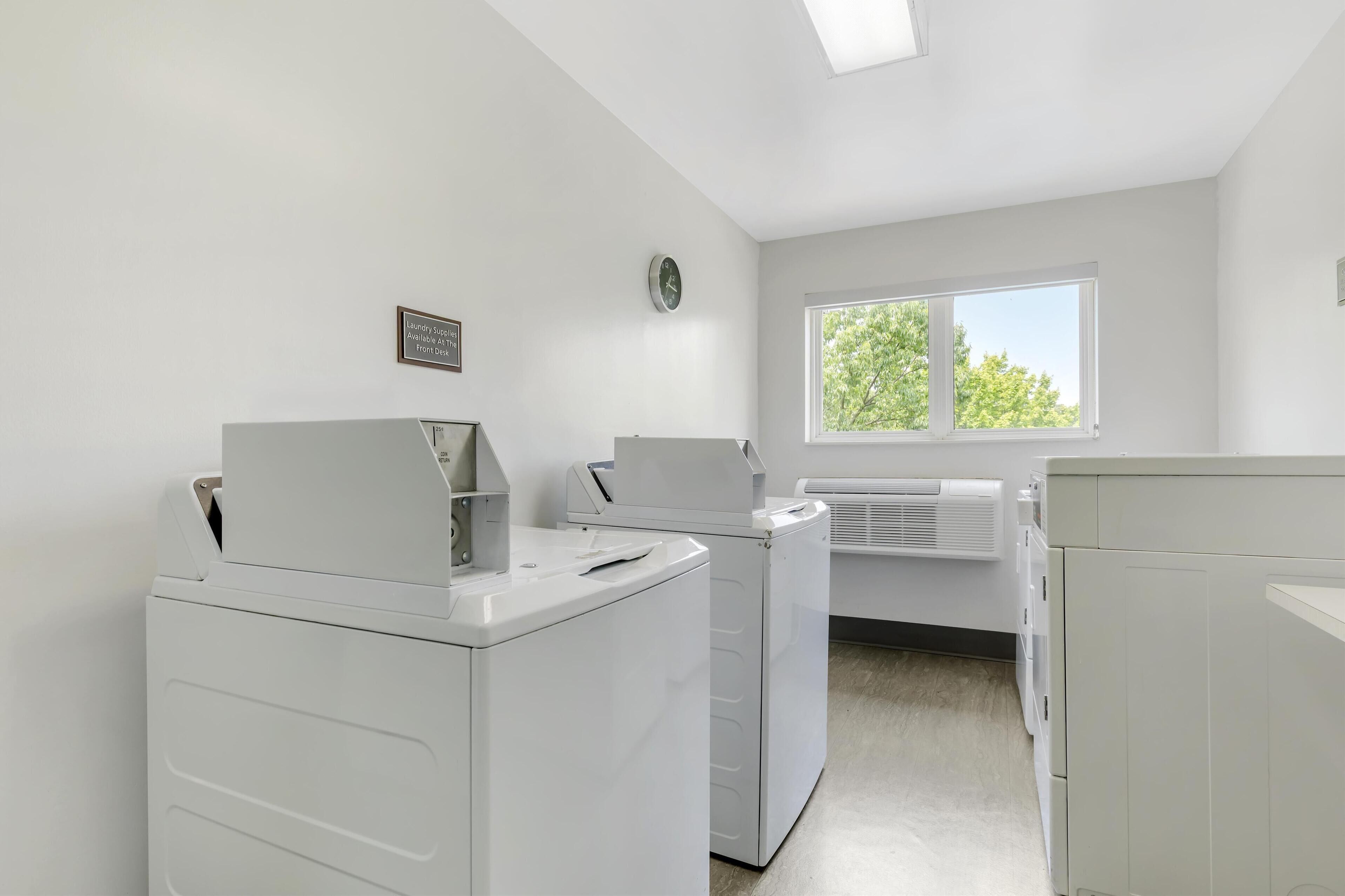 A laundry room with white appliances, a clock, and a window with a view of trees.
