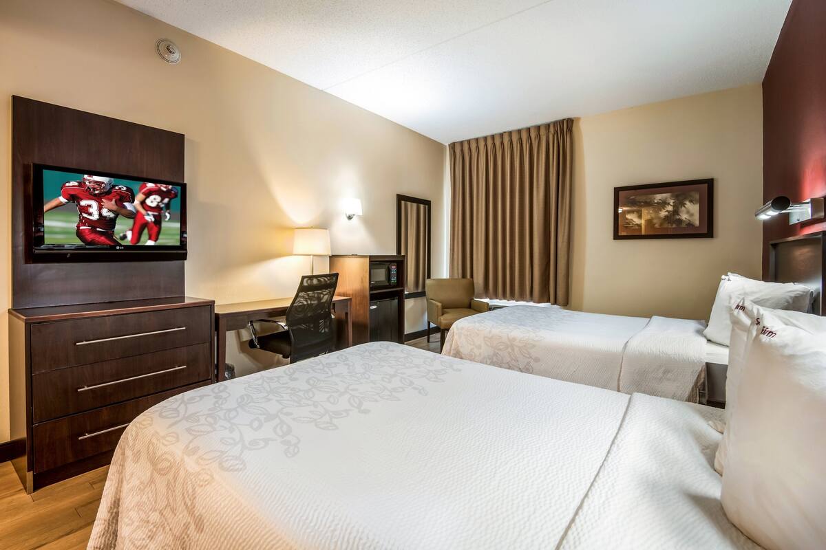 A hotel room with two beds, a TV on the wall, a desk with a chair, a lamp, and a painting on the wall.