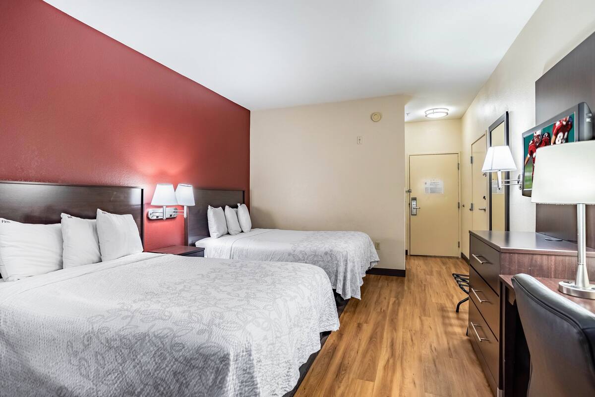 A hotel room with two beds, a wooden floor, a red accent wall, a desk with a lamp, and a mirror.