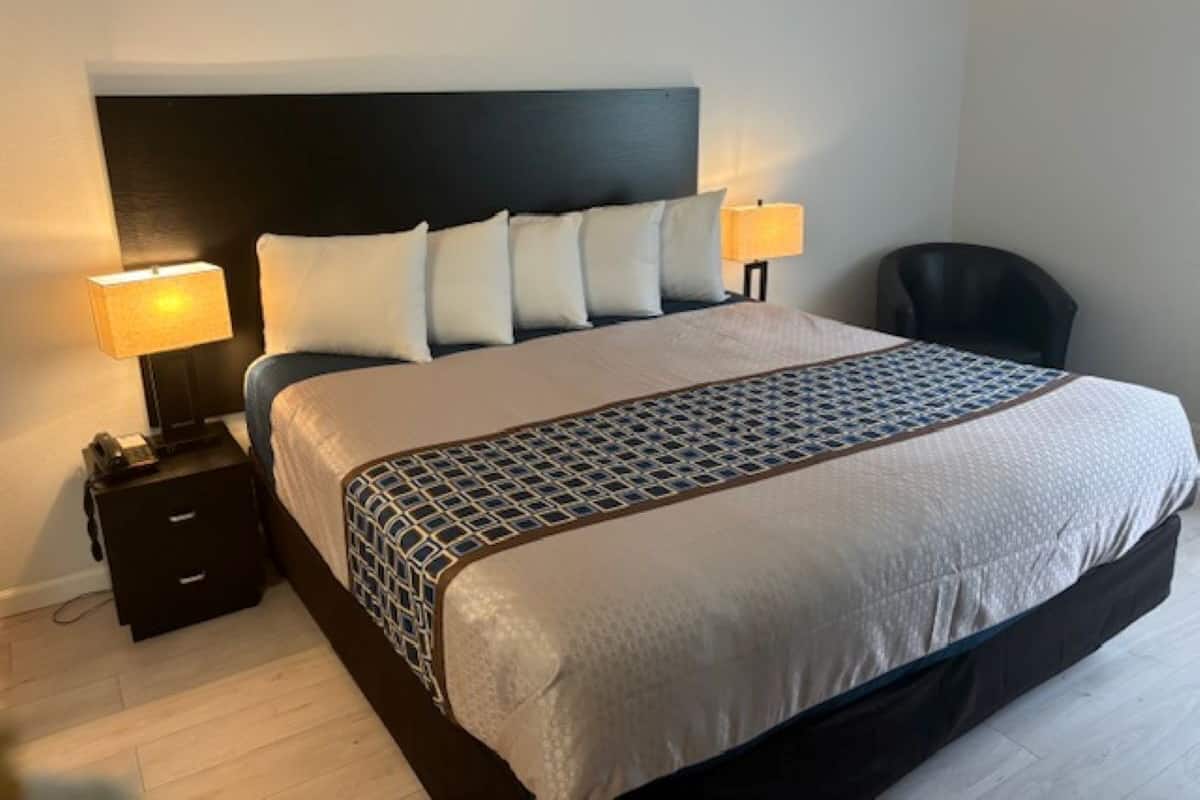 A neatly made bed with a patterned bedspread, two bedside lamps, a small drawer, and a chair in a hotel room.