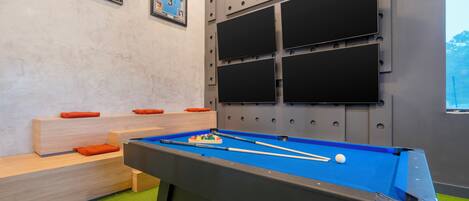 A pool table with balls and cues, a sofa with orange cushions, and two framed sports jerseys on the wall.