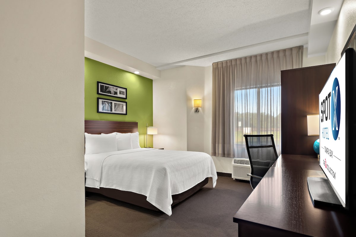 Habitación de hotel con cama, escritorio, silla, l ámpara y ventana con cortinas.