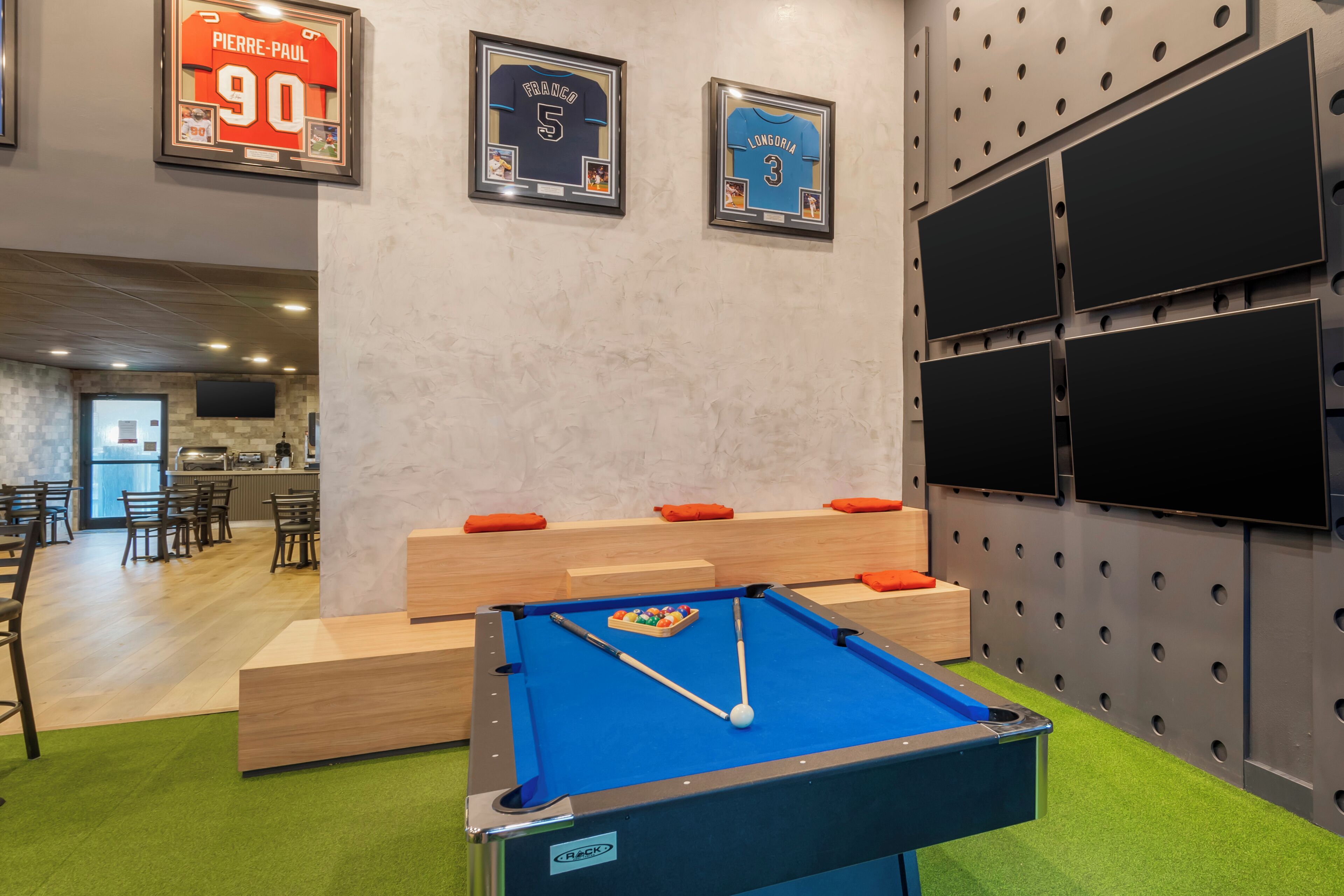 Une table de billard avec un queue et des billes, un coin salon avec des coussins orange et, au mur, des maillots de sport encadrés.