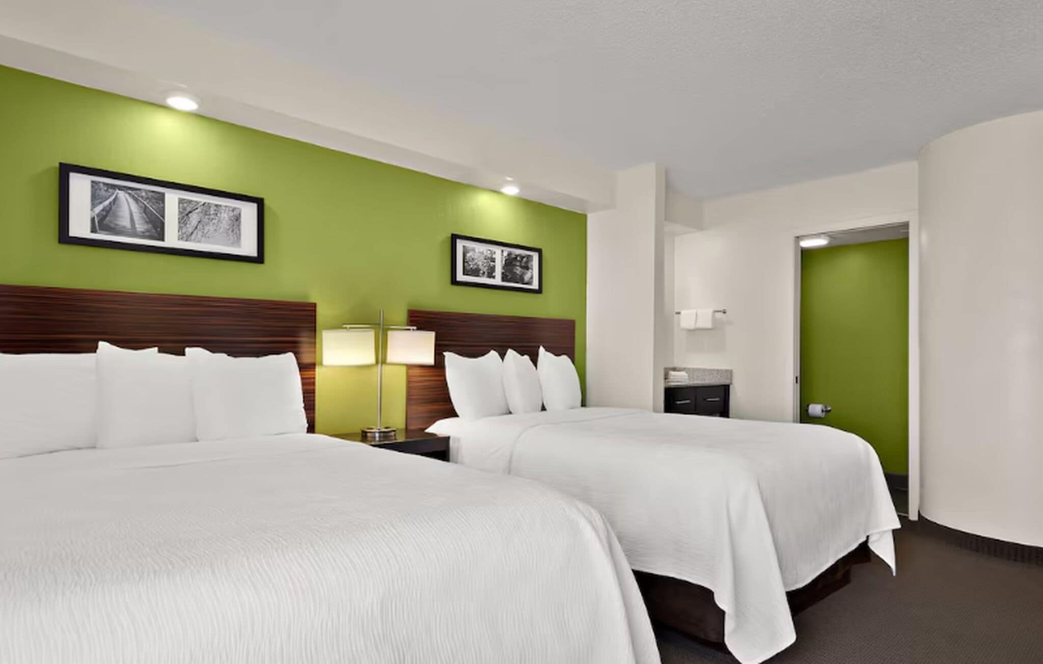 Habitación de hotel con dos camas, paredes verdes, cuadros enmarcados, una mesita de noche con lámpara y la puerta del baño.