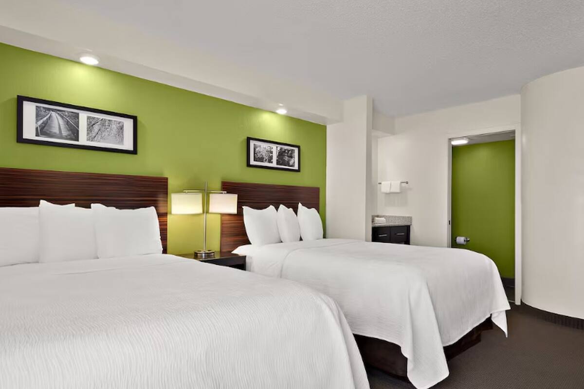 A hotel room with two beds, green walls, framed pictures, a nightstand with a lamp, and a bathroom door.