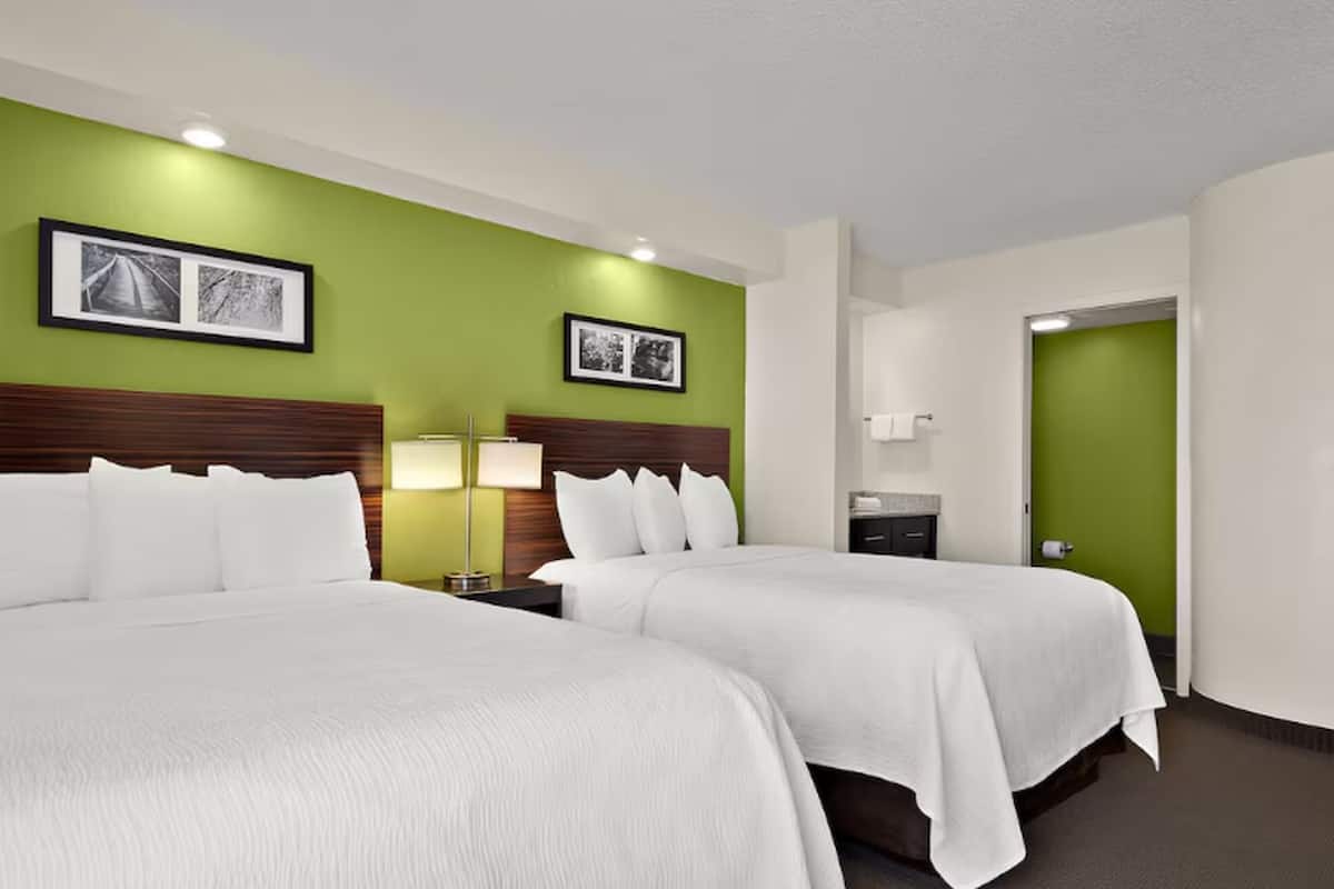 Habitación de hotel con dos camas, paredes verdes, cuadros enmarcados, una mesita de noche con lámpara y la puerta del baño.