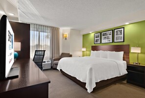 Habitación de hotel con cama, escritorio, silla, ventana con cortinas y marcos de cuadros en la pared.