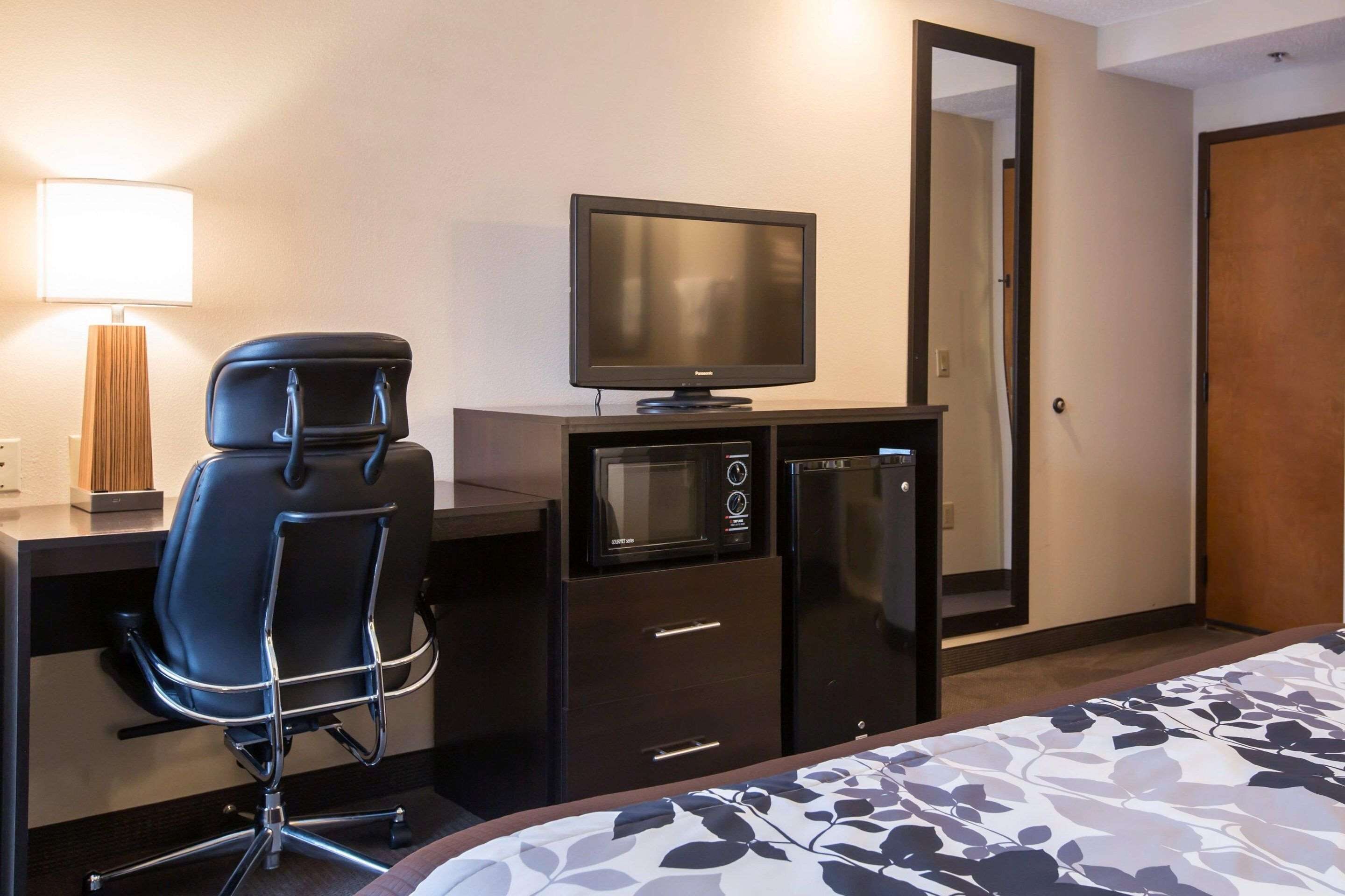 A hotel room with a bed, a desk with a computer, a chair, a television, a microwave, a refrigerator, and a mirror.