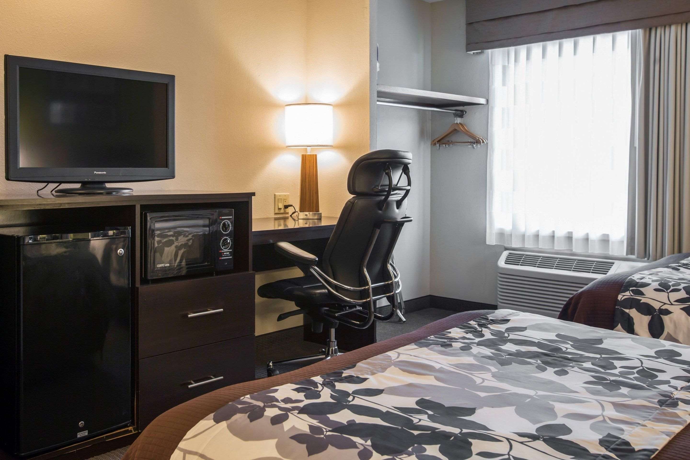 A hotel room with a bed, a desk with a chair, a television, a mini-fridge, a lamp, and a window with curtains.