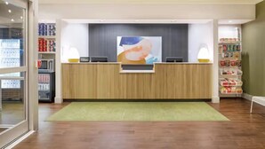 A reception desk with a green rug, a large abstract painting, and shelves with various items. - Spark by Hilton Grand Rapids (Grand Rapids)