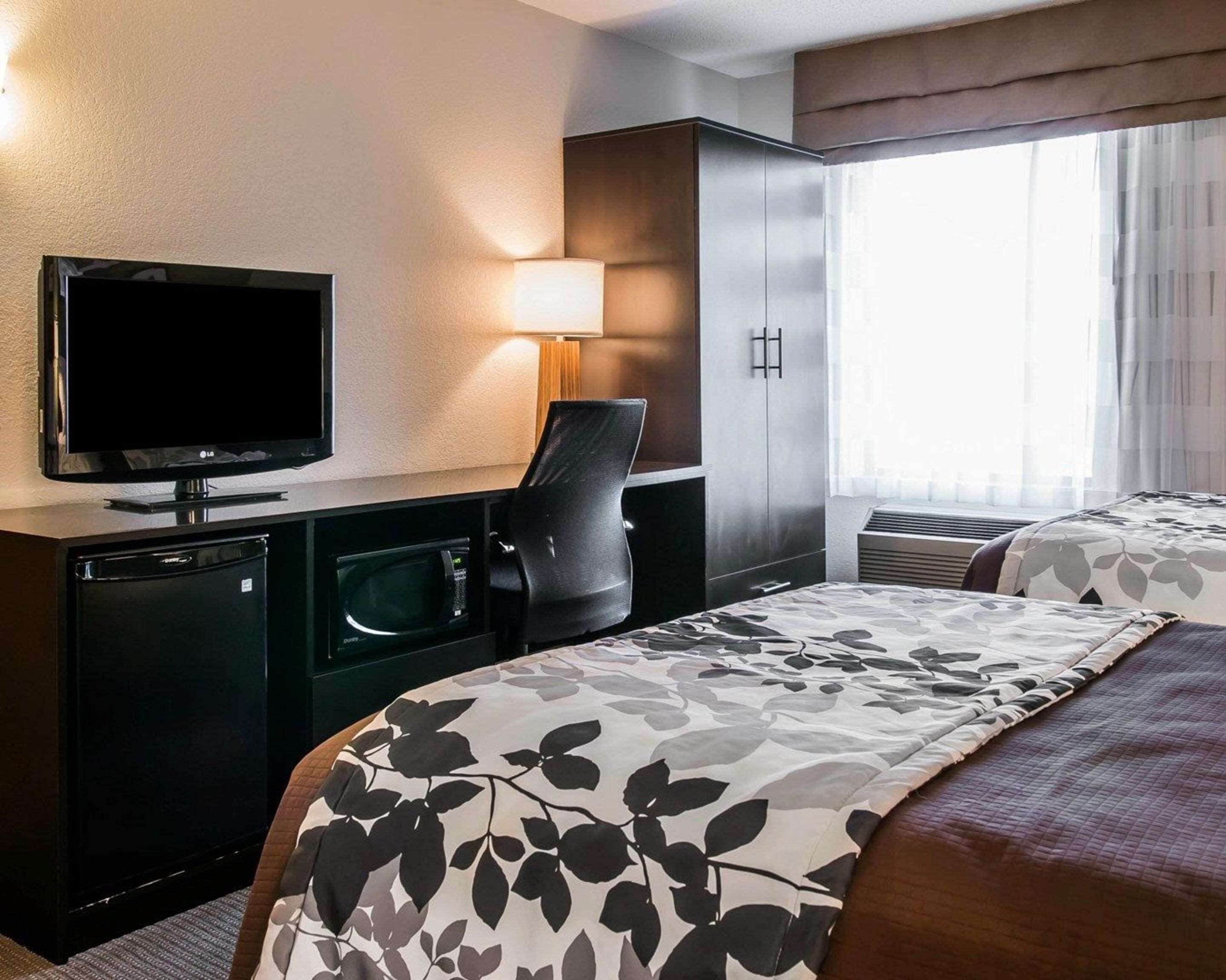 Habitación de hotel con televisión de pantalla plana, escritorio con lámpara, microondas, refrigerador pequeño y dos camas con cubrecamas estampadas con motivos florales.