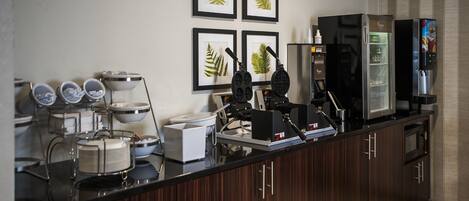 A modern kitchen with a black countertop, a coffee machine, a refrigerator, and framed leaf artwork on the wall.