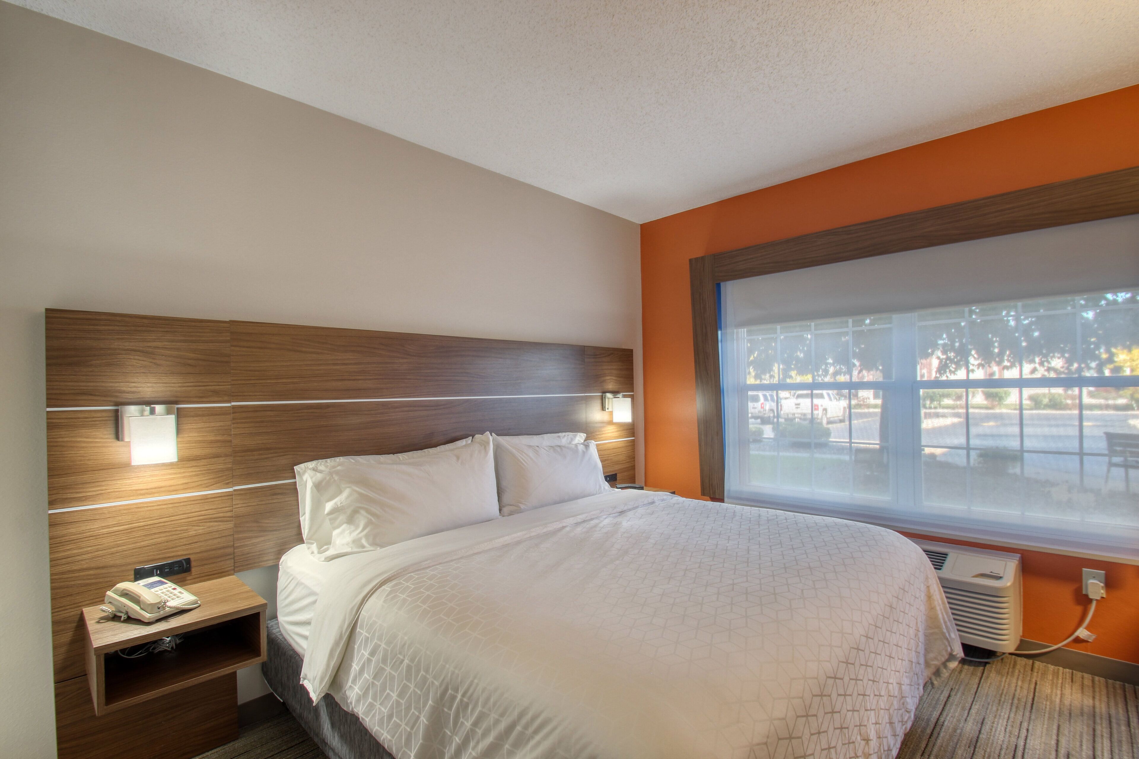 Une chambre d’hôtel avec une tête de lit en bois, un grand lit avec une literie blanche, une table de chevet avec un téléphone et une fenêtre donnant sur l’extérieur.