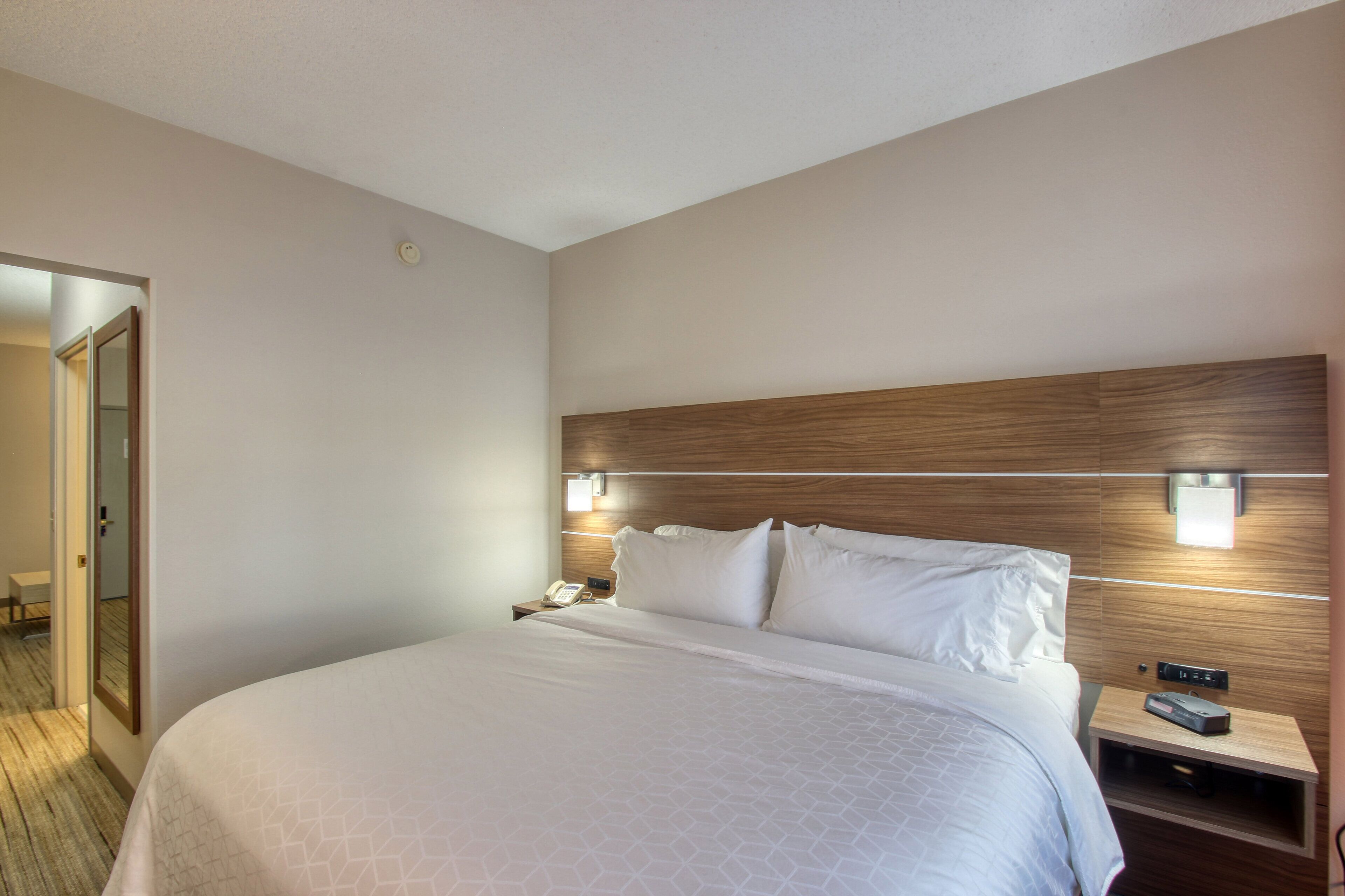 A hotel room with a large bed, a wooden headboard, bedside lamps, a nightstand with a phone and a remote control, and a door leading to another room.