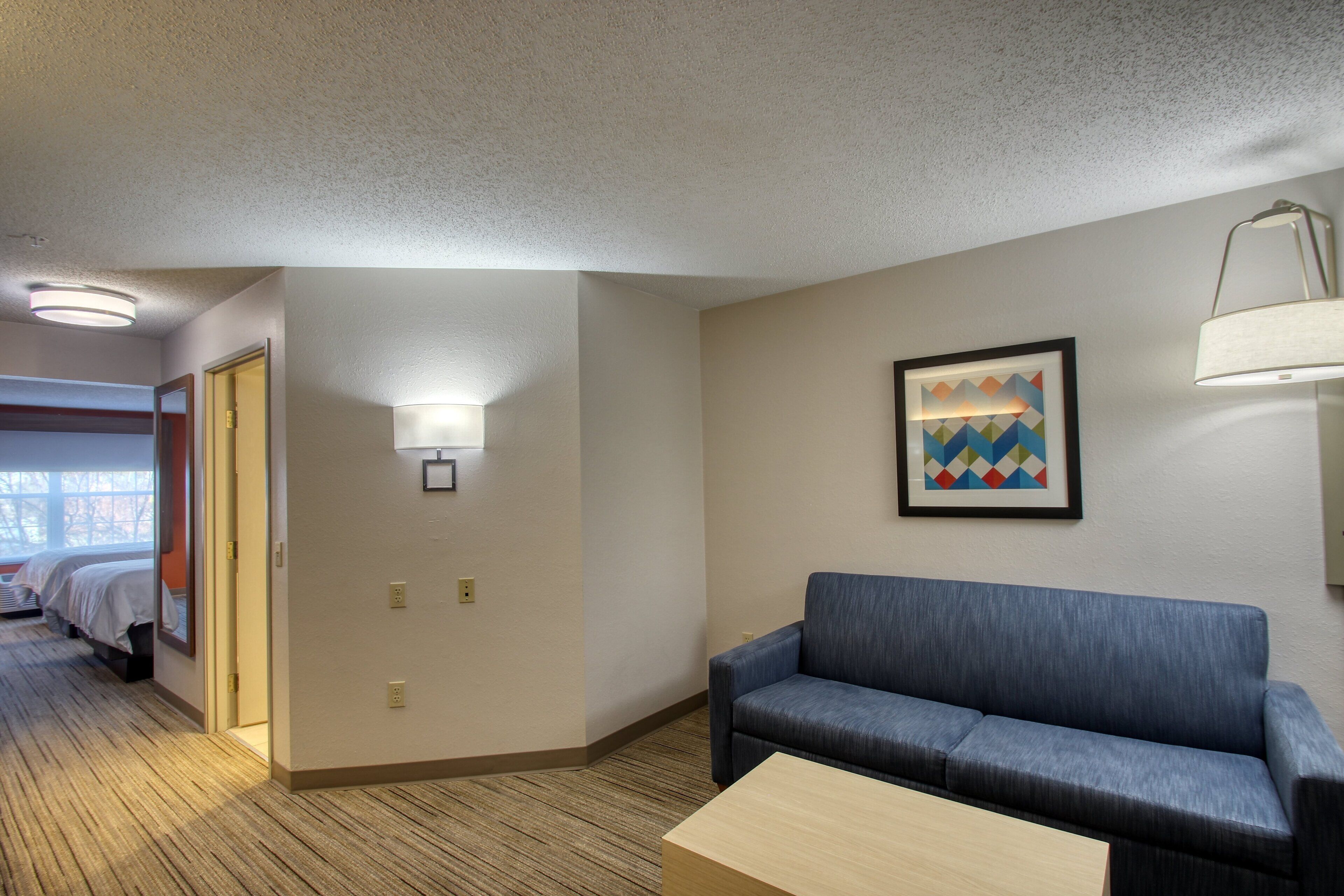 A hotel room with a blue sofa, a wooden coffee table, a framed picture on the wall, and a door leading to another room.