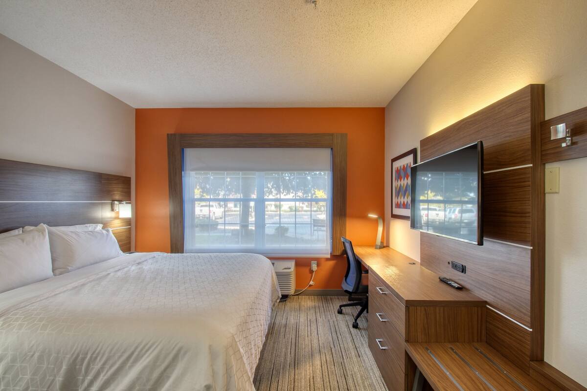 A hotel room with a large bed, a desk with a computer, a television, and a window with a view of trees.