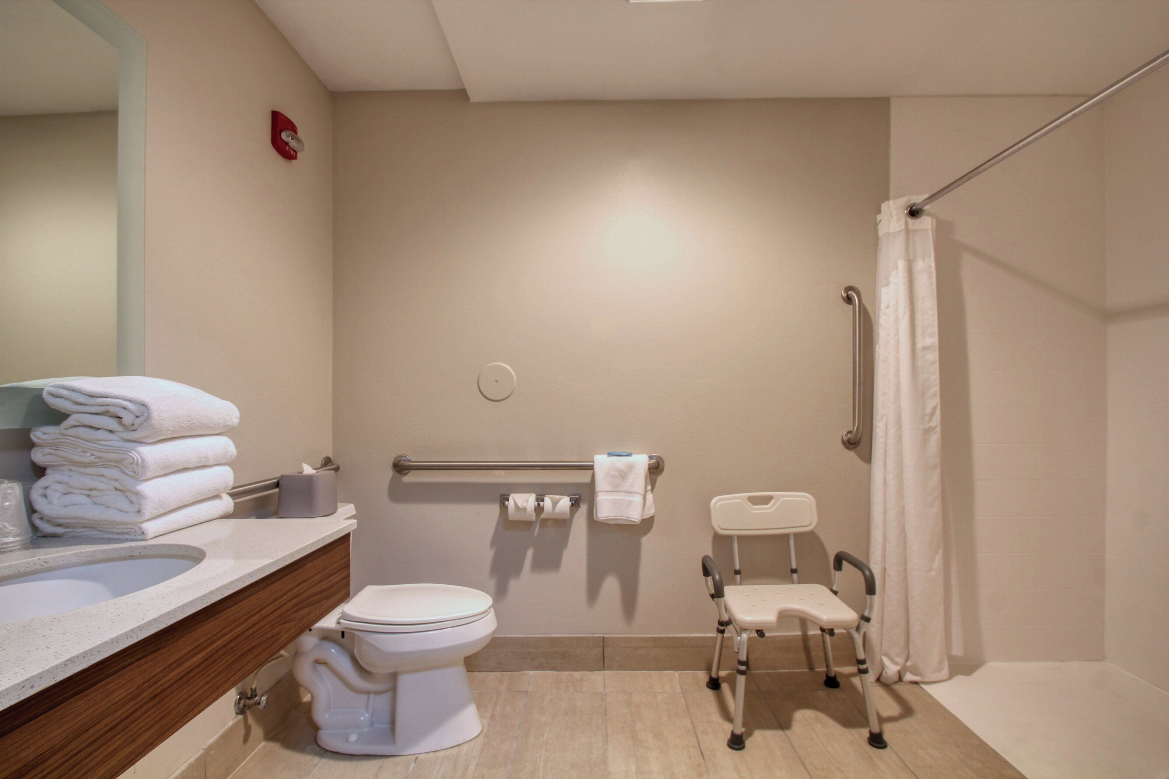 A bathroom with a toilet, a sink, a mirror, a towel rack, a grab bar, a shower curtain, and a wheelchair-accessible bench.