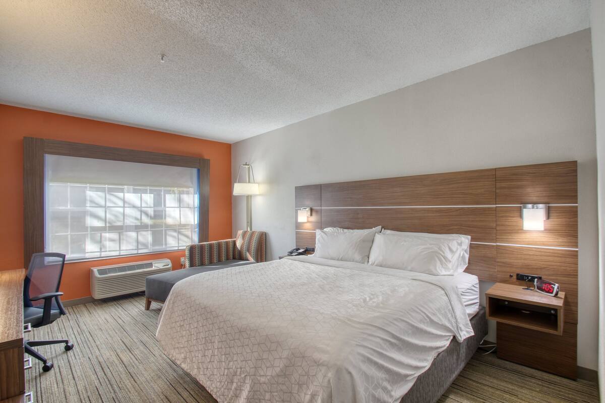 A hotel room with a large bed, a desk, a chair, and a window with blinds.