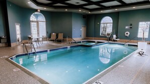 Uma piscina interior com água cristalina, bordas azulejadas e uma escada de acesso. Há espreguiçadeiras e uma pequena mesa perto da piscina.