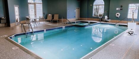 Una piscina interior con agua cristalina, bordes azulejados y una escalera de piscina. Hay sillones de descanso y una pequeña mesa cerca de la piscina.