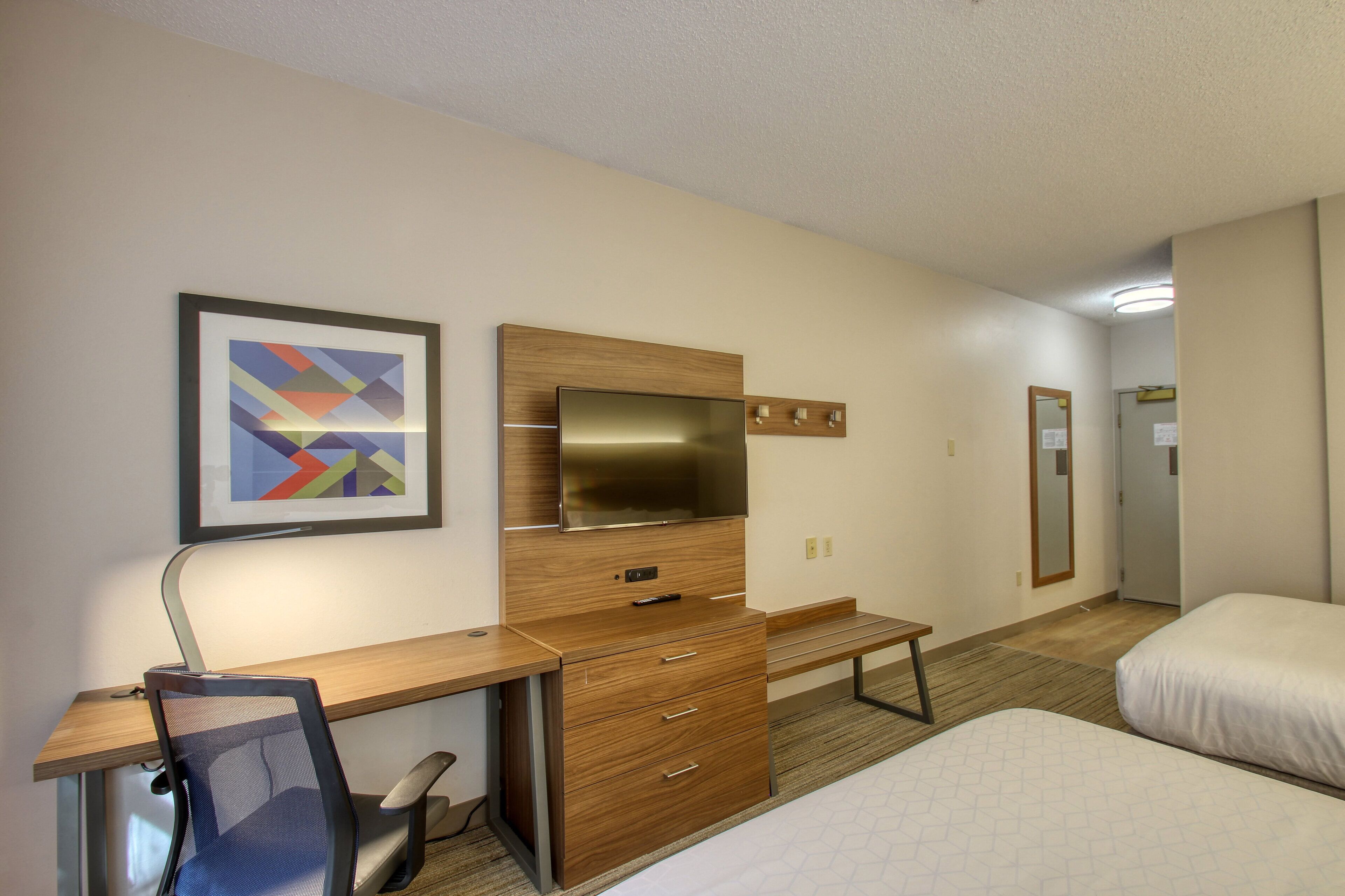 A hotel room with a wooden desk, a television, a bed, and a framed picture on the wall.