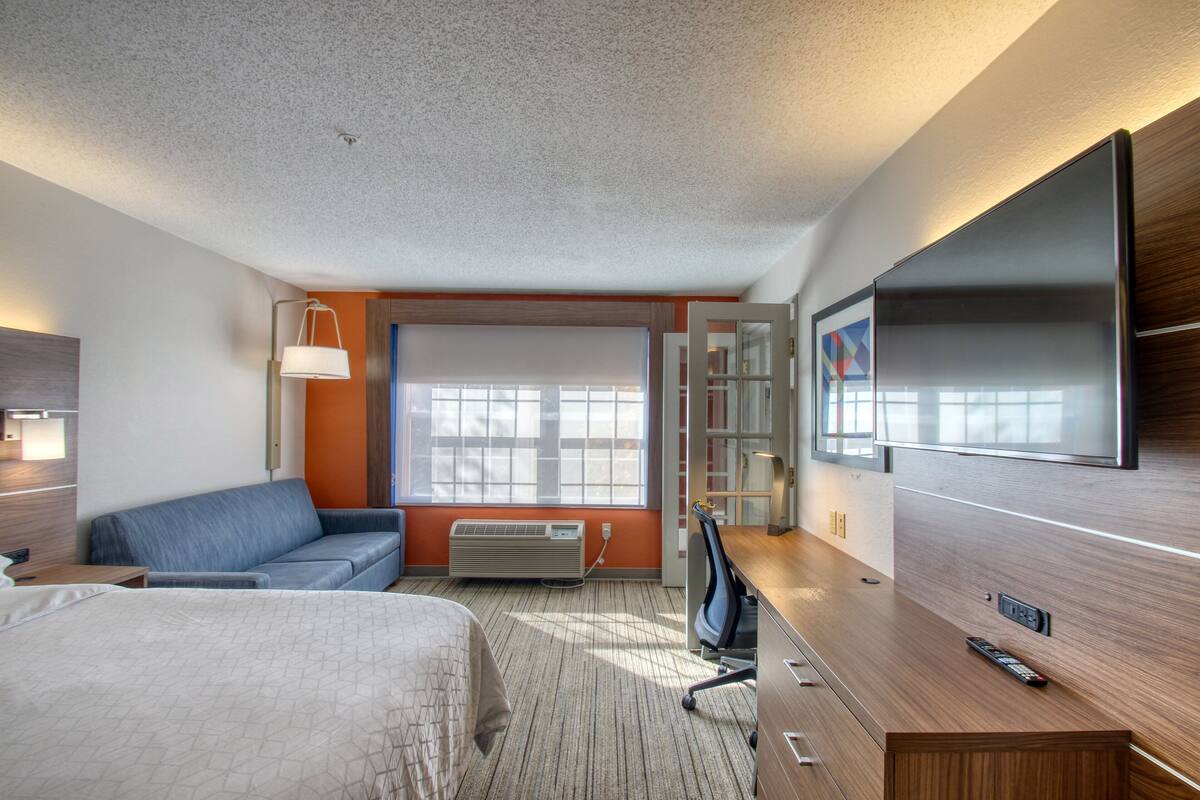 A hotel room with a bed, a sofa, a desk, a TV, and a window with blinds.
