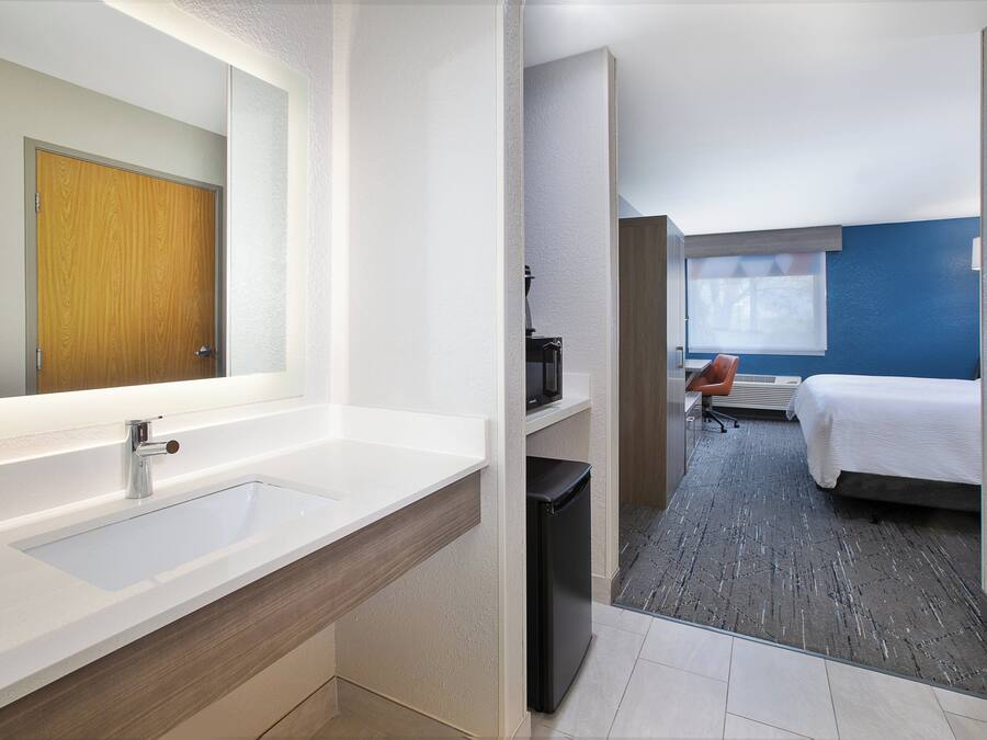 A modern hotel room with a large bathroom sink, a compact refrigerator, a bed with a white cover, and a blue accent wall.