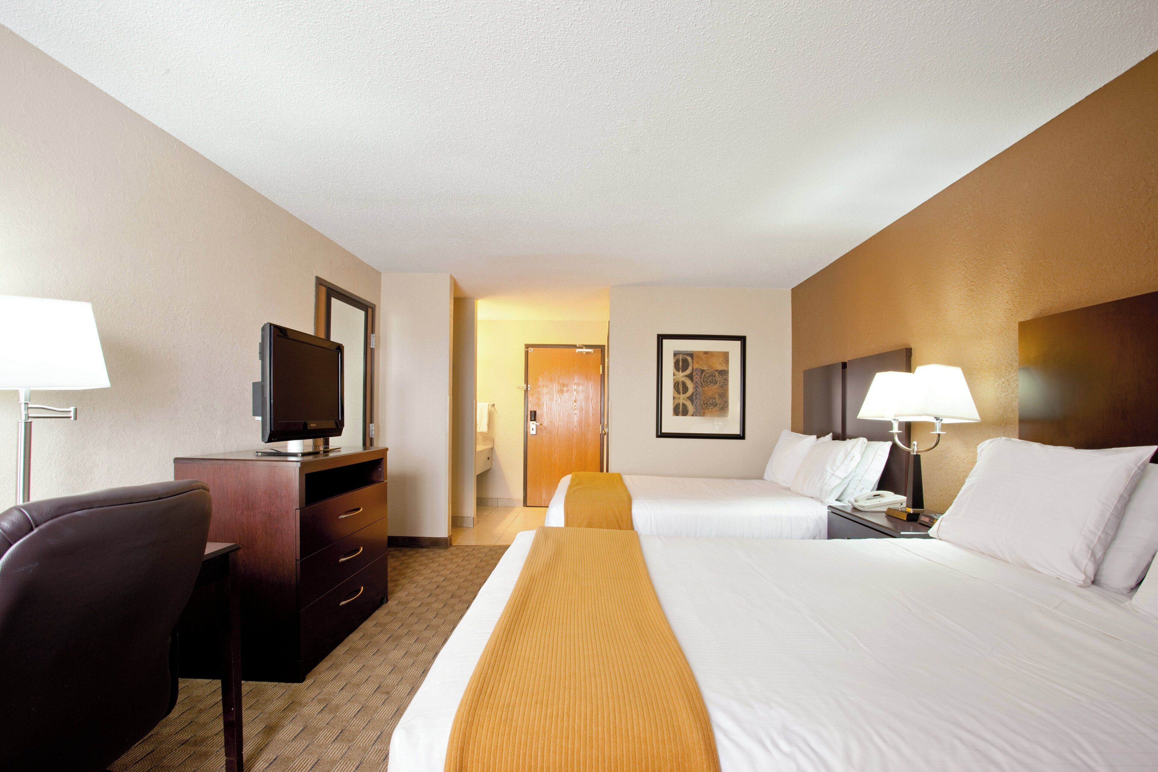 A hotel room with two beds, a desk with a television, a chair, a bedside table with a lamp, and a painting on the wall.
