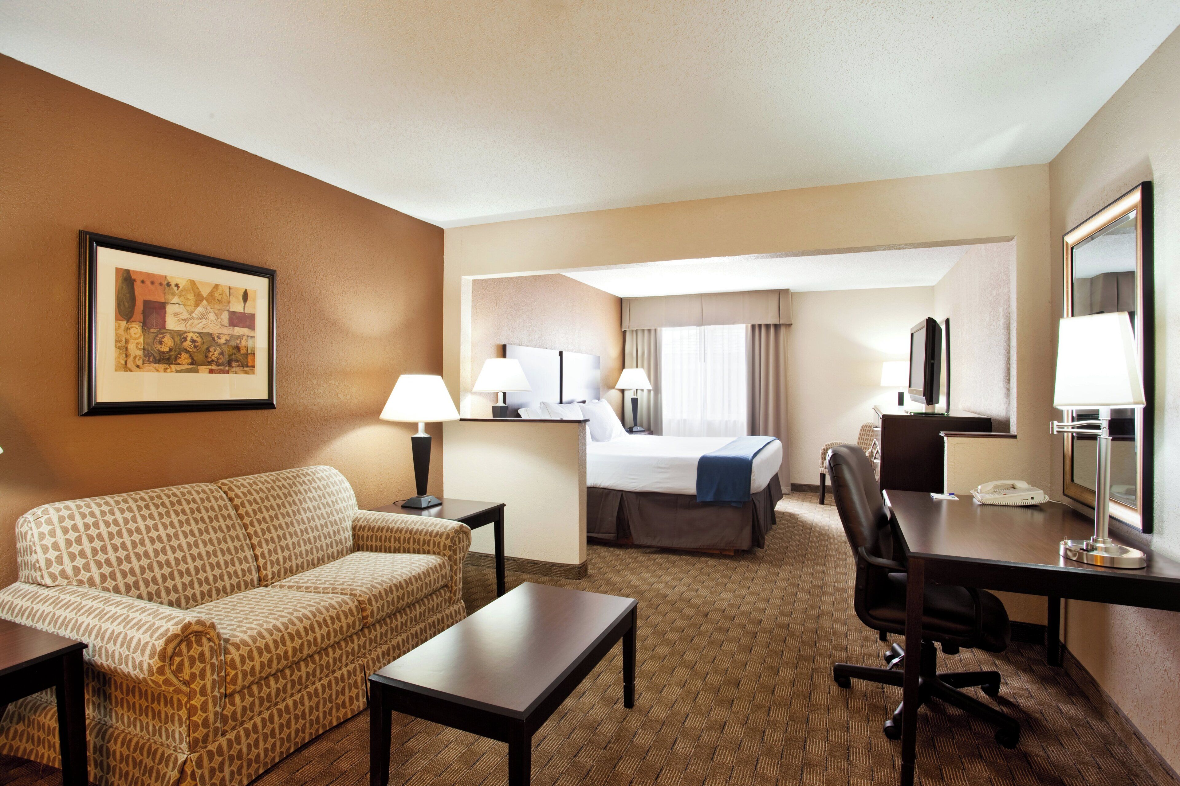 A hotel room with a sofa, a desk, a chair, a bed, and a desk lamp.