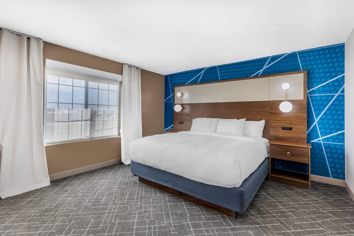 A hotel room with a large bed, a nightstand, a mirror with lights, and a window with curtains.