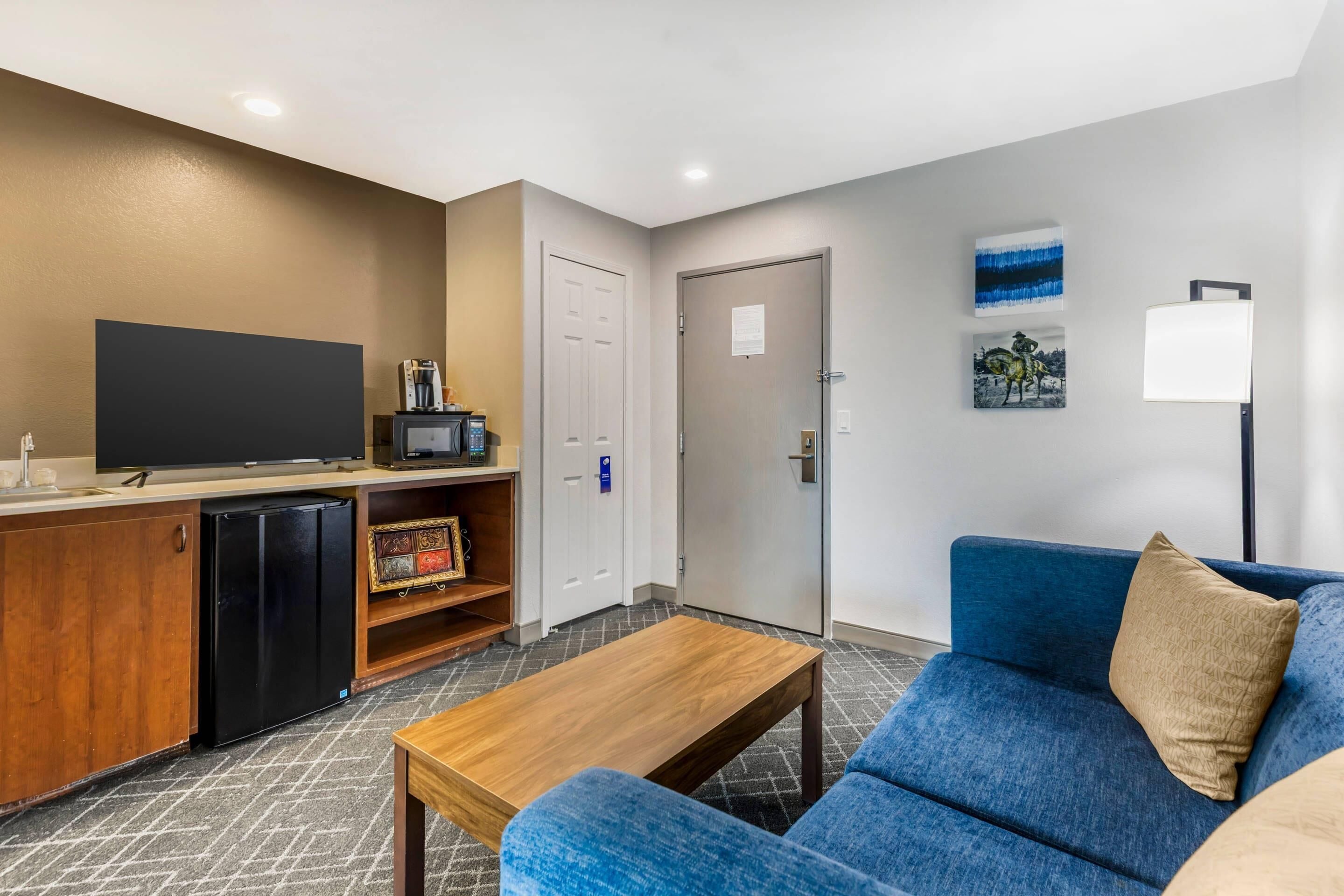 A modern living room with a blue sofa, a wooden coffee table, a flat-screen TV, a mini-fridge, and a small kitchen area with a microwave.