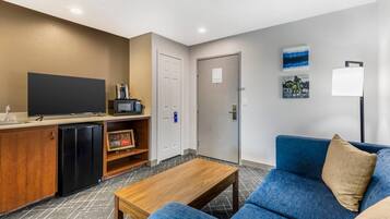 A modern living room with a blue sofa, a wooden coffee table, a flat-screen TV, a mini-fridge, and a small kitchen area with a microwave.