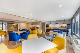Un comedor moderno con sillas amarillas y azules, una mesa central y una cocina al fondo.