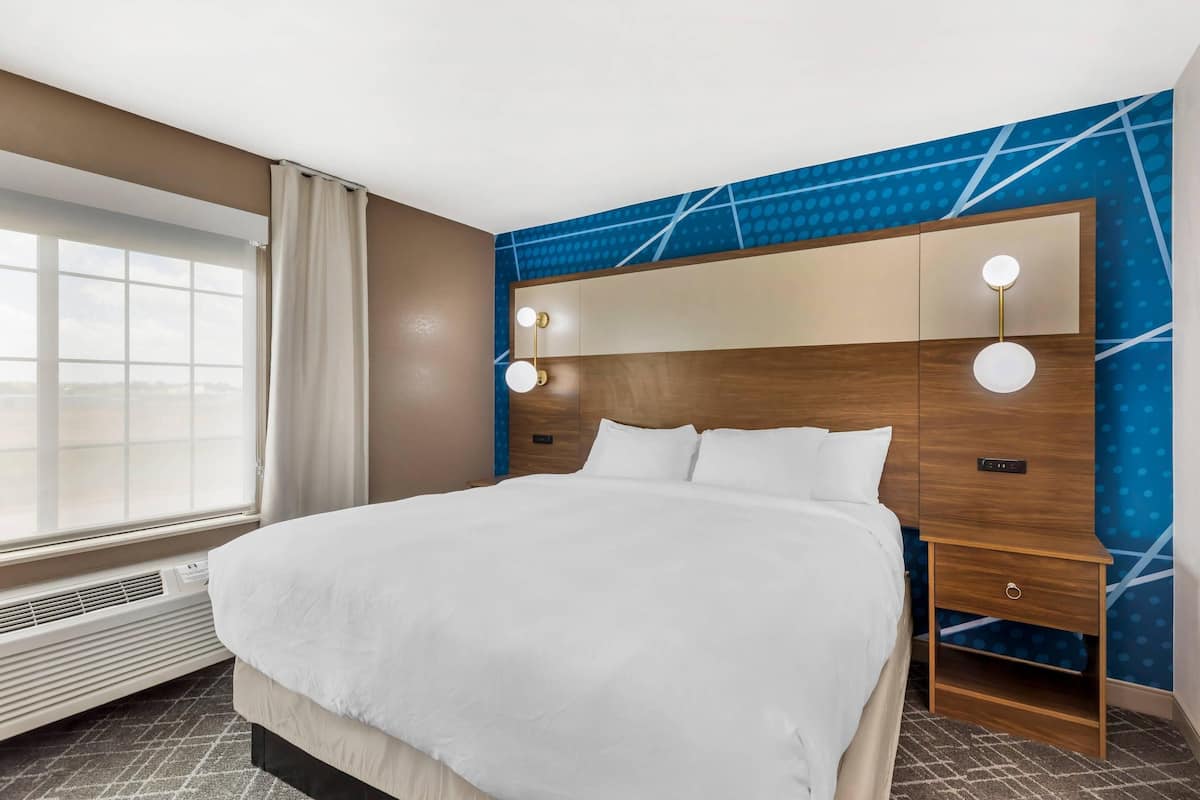 A hotel room with a large bed, a nightstand, a wall-mounted lamp, and a window with curtains.