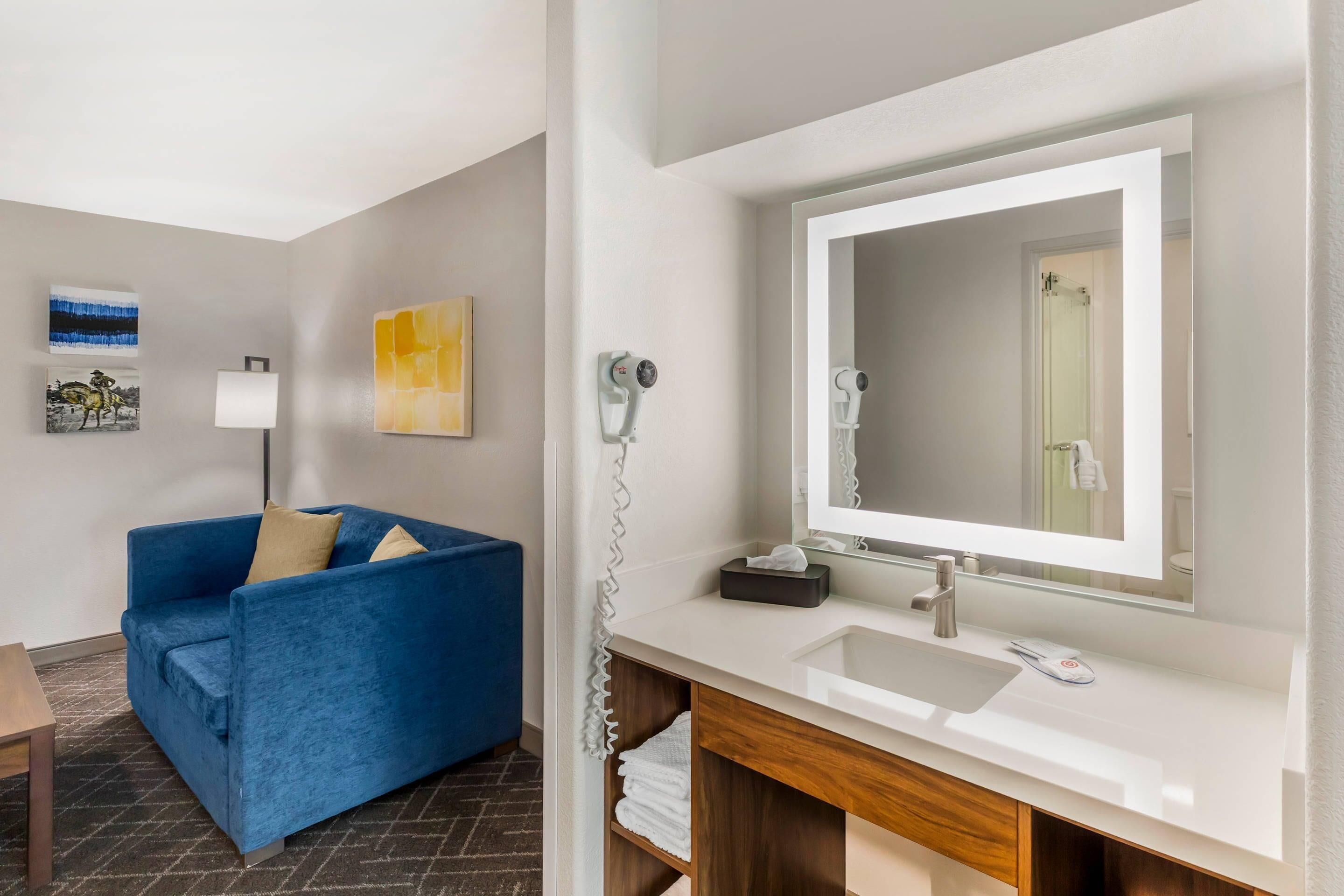 Une salle de bain d’hôtel avec un lavabo blanc, un grand miroir, un sèche-cheveux et un canapé bleu dans la pièce adjacente.