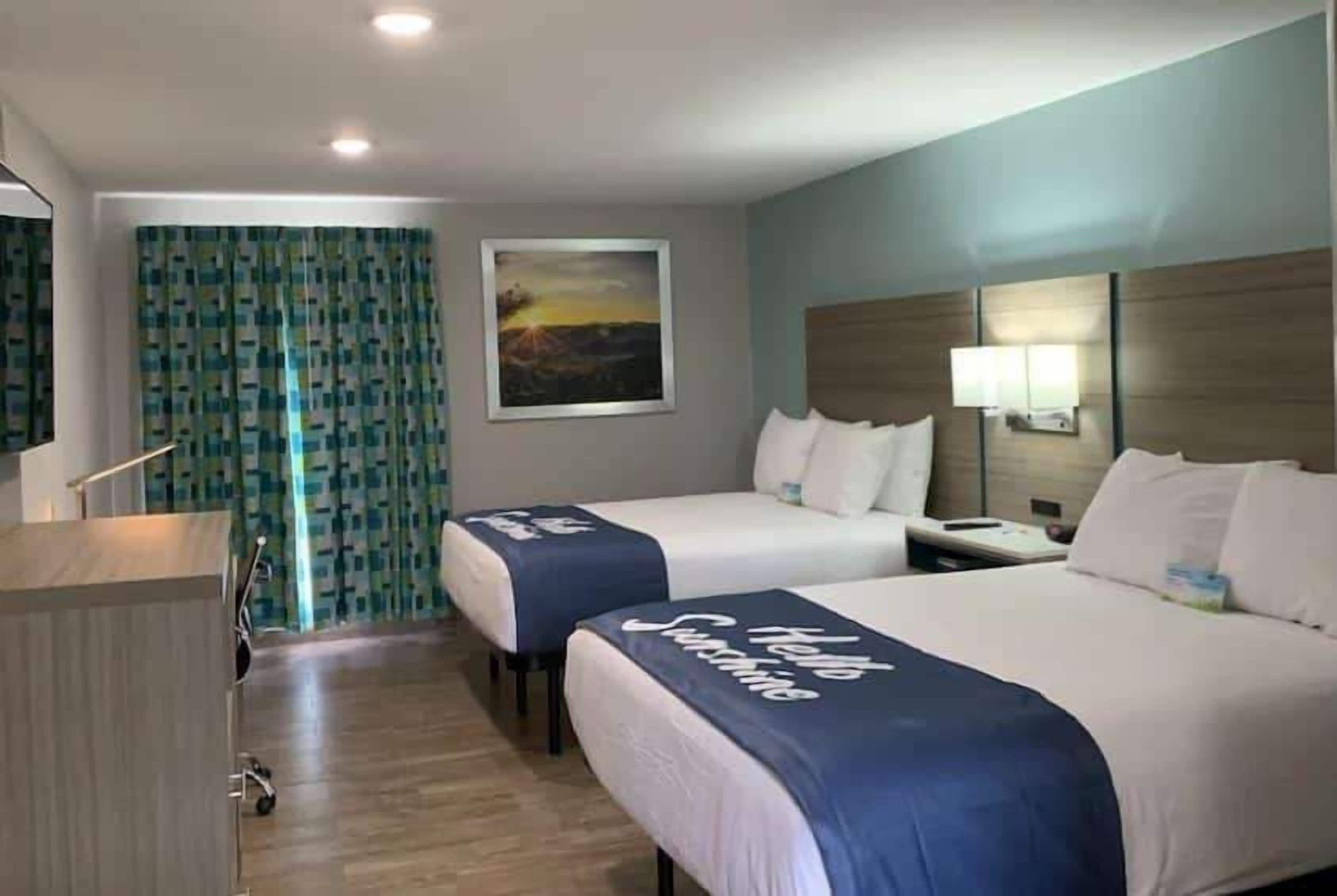 Habitación de hotel con dos camas, un escritorio, una silla, un ventanal con cortinas y un cuadro en la pared.