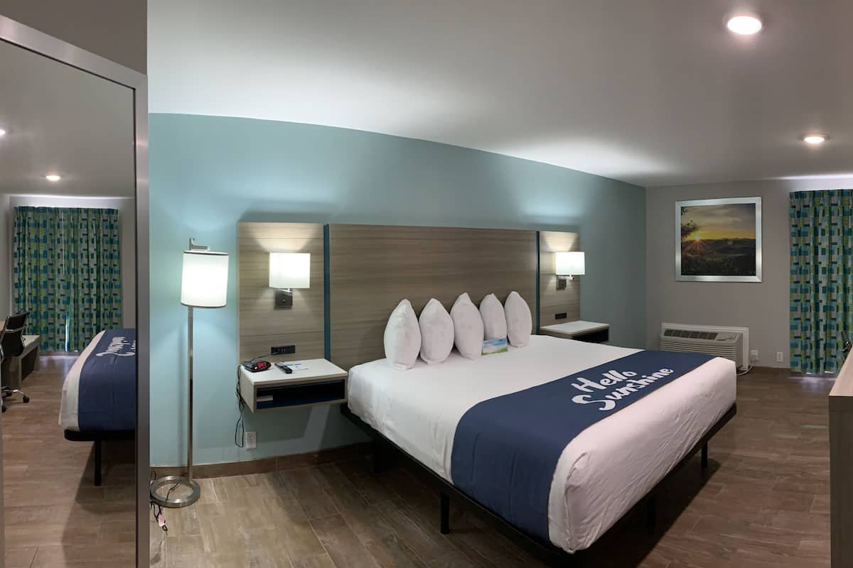 Ein Hotelzimmer mit einem großen Bett, zwei Nachttischen, einem Schreibtisch, einem Stuhl, einem Spiegel, einem Fenster mit Vorhängen und einem am Wand montierten Fernseher.