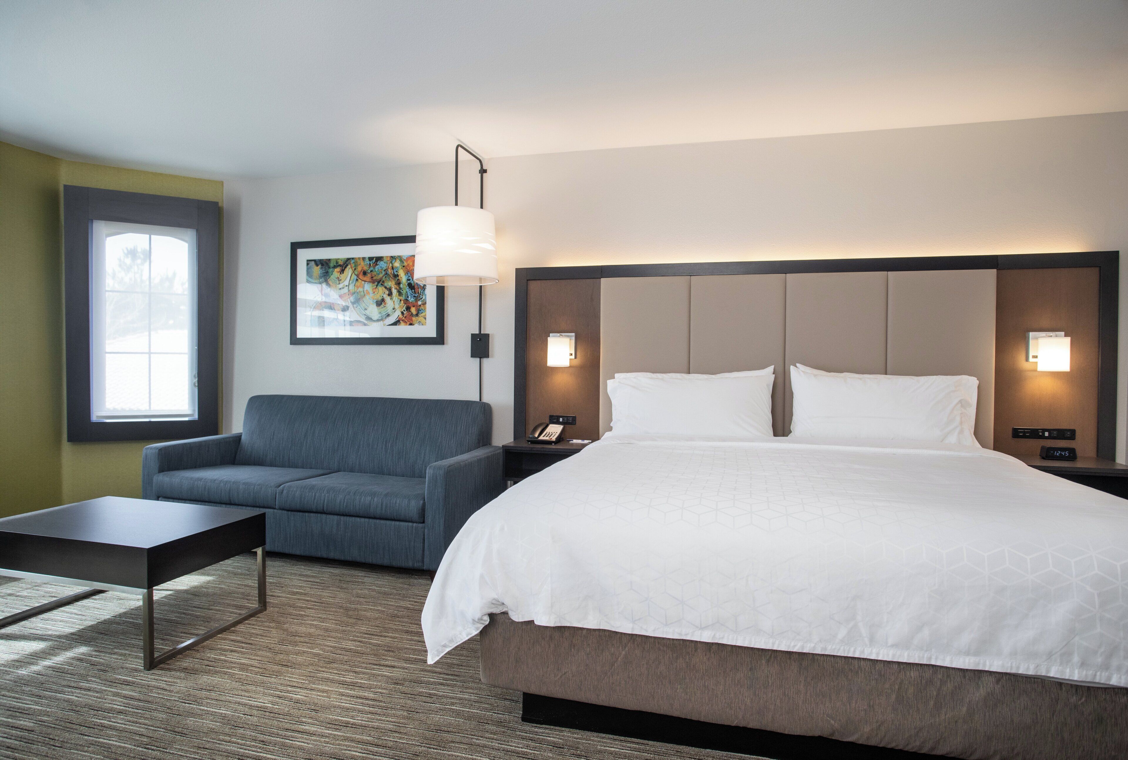 A hotel room with a bed, a sofa, a coffee table, a painting on the wall, and two bedside lamps.