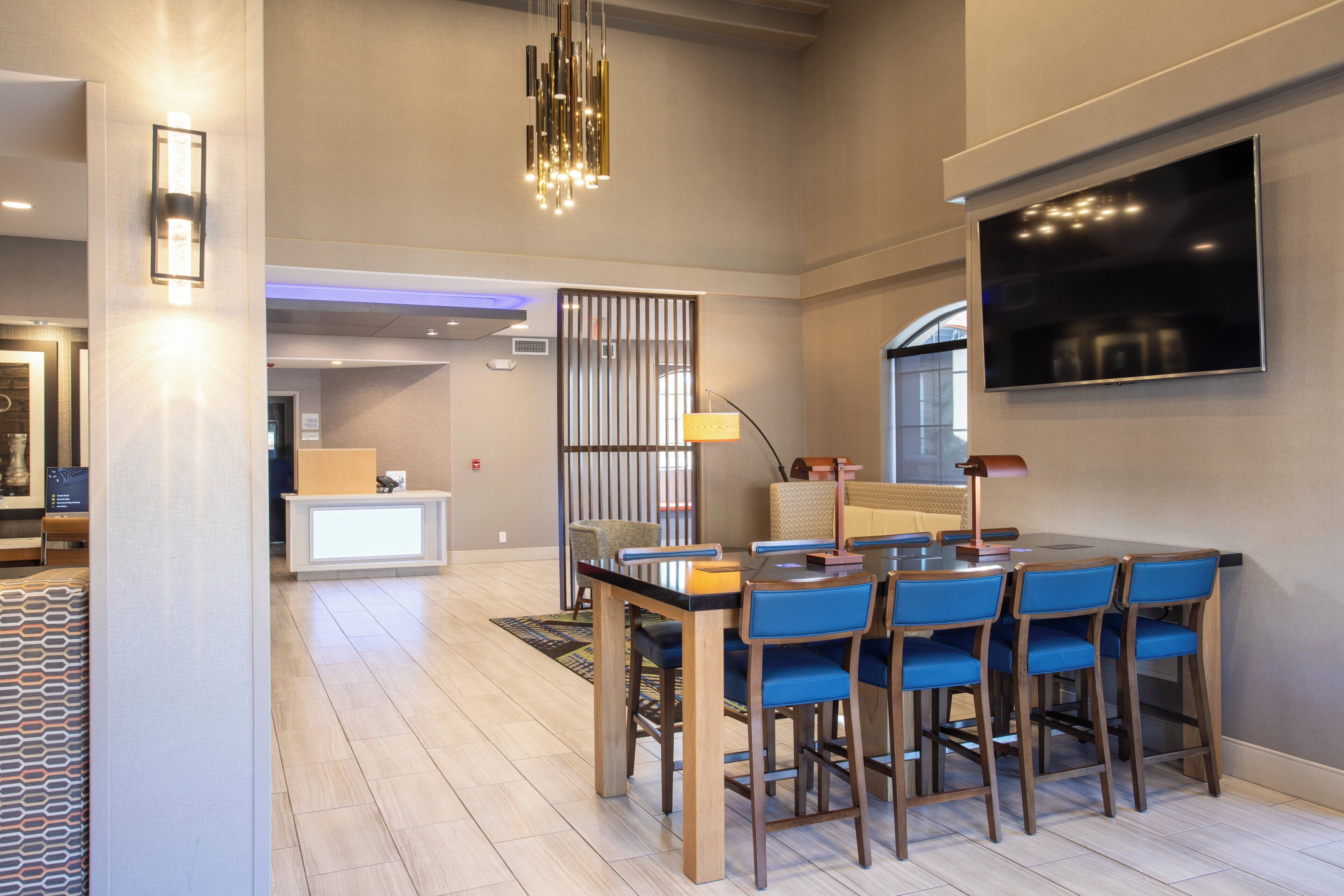 A modern dining area with a long table and blue chairs, a TV mounted on the wall, and a patterned rug on the floor.