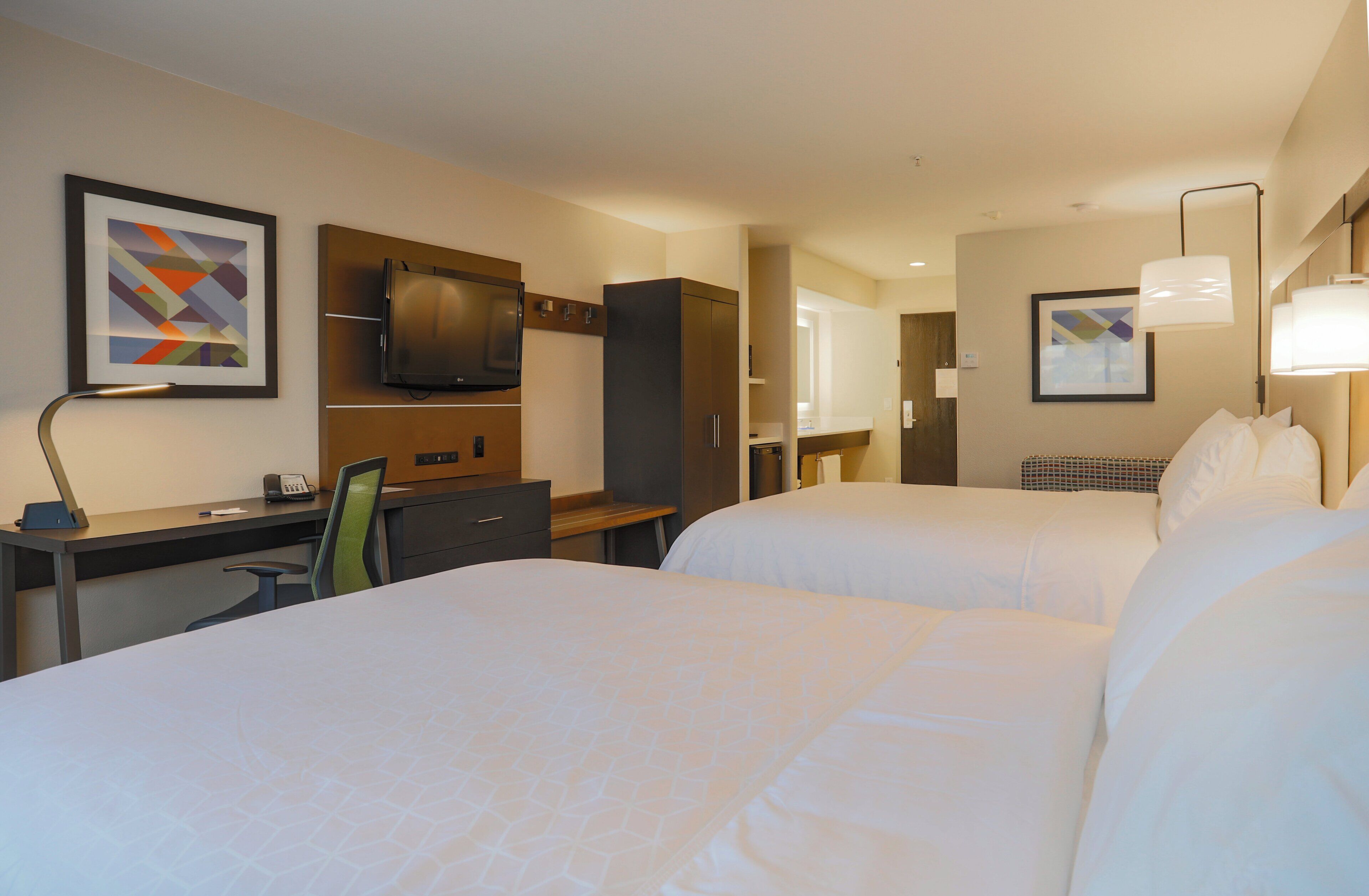 A hotel room with two beds, a desk, a chair, a TV, and artwork on the walls.