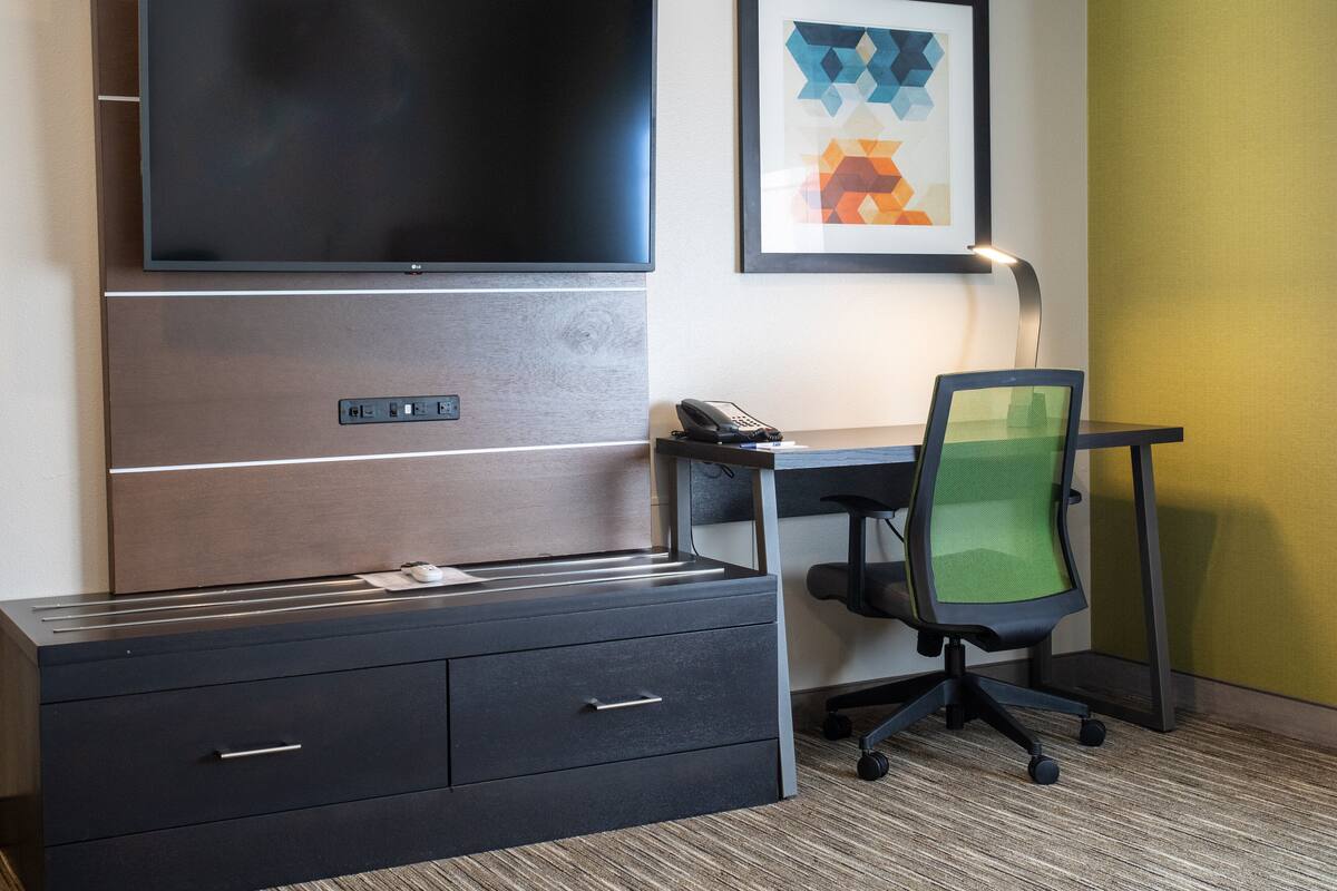 A hotel room with a flat-screen TV mounted on the wall, a desk with a chair, and a framed abstract artwork above the desk.