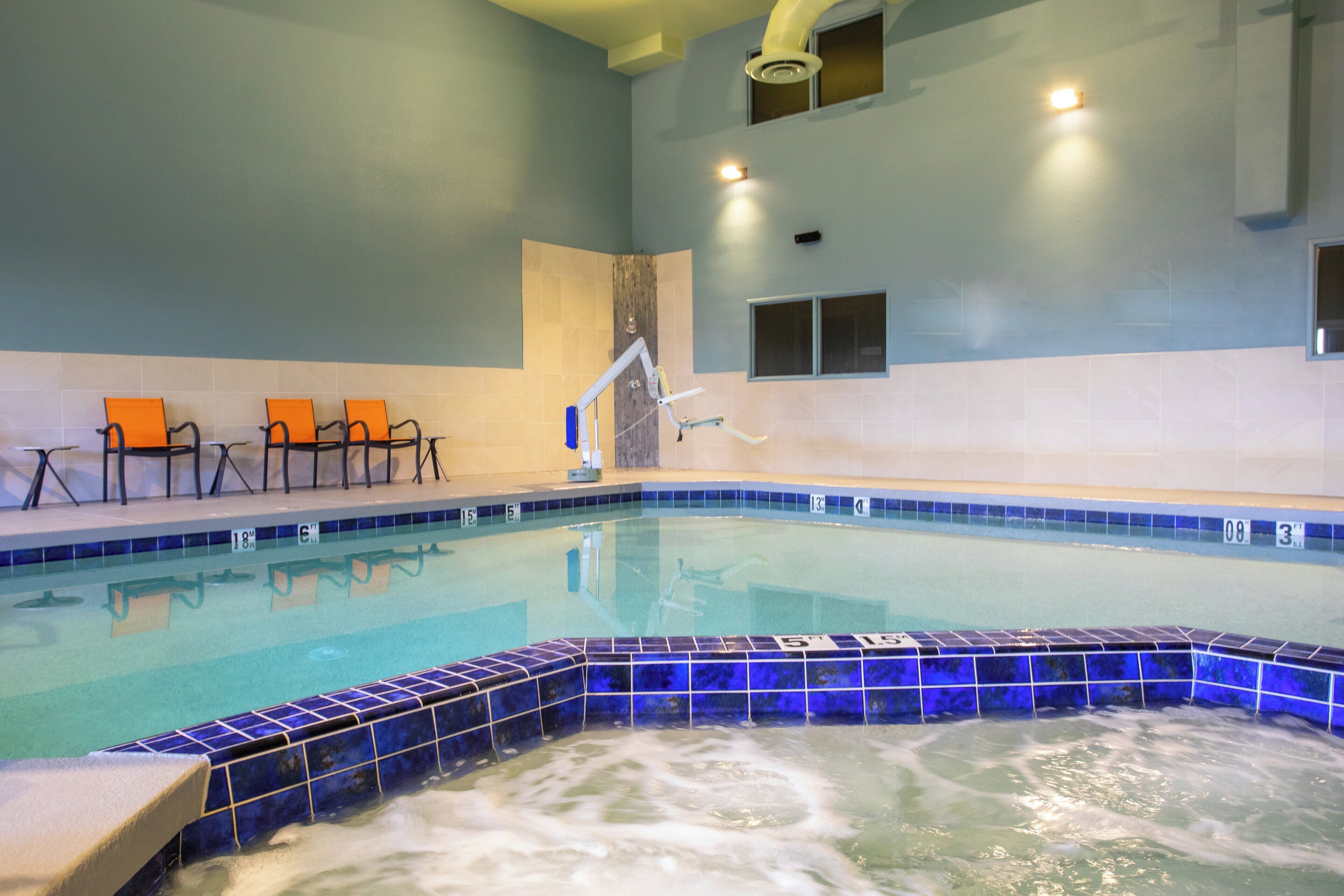 An indoor swimming pool with blue-tiled edges, a hot tub area, and orange chairs along the wall.
