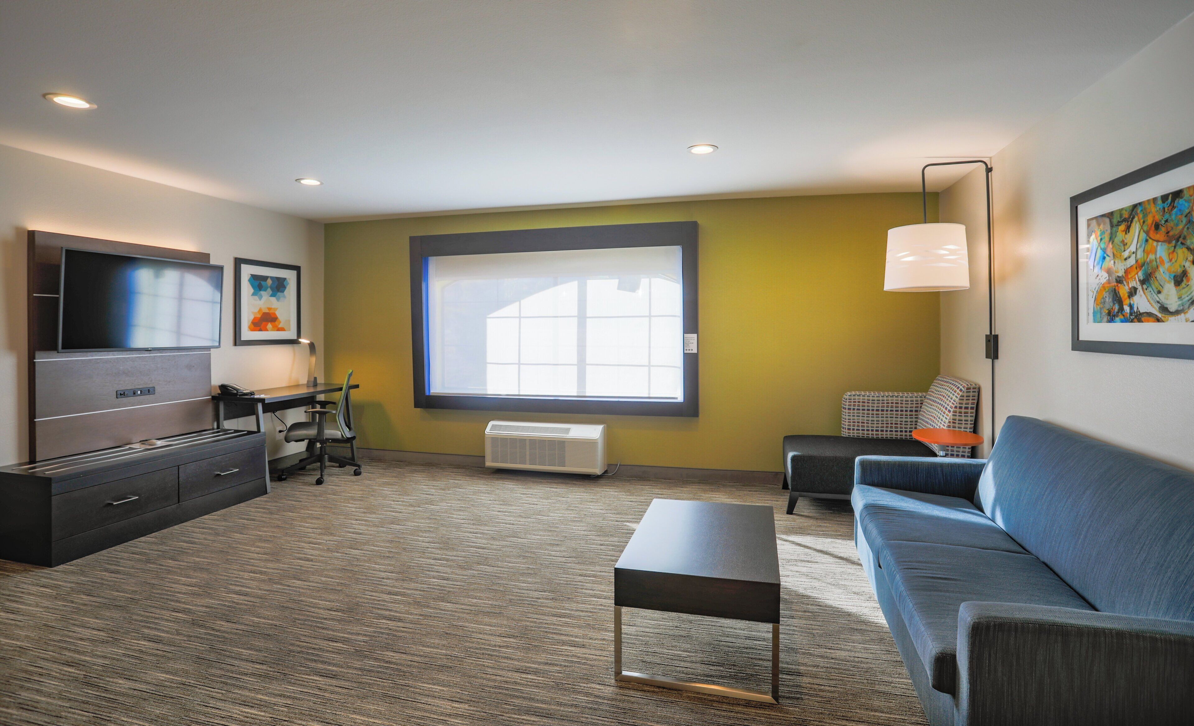 A modern hotel room with a large window, a flat-screen TV, a sofa, a desk, and a lamp.