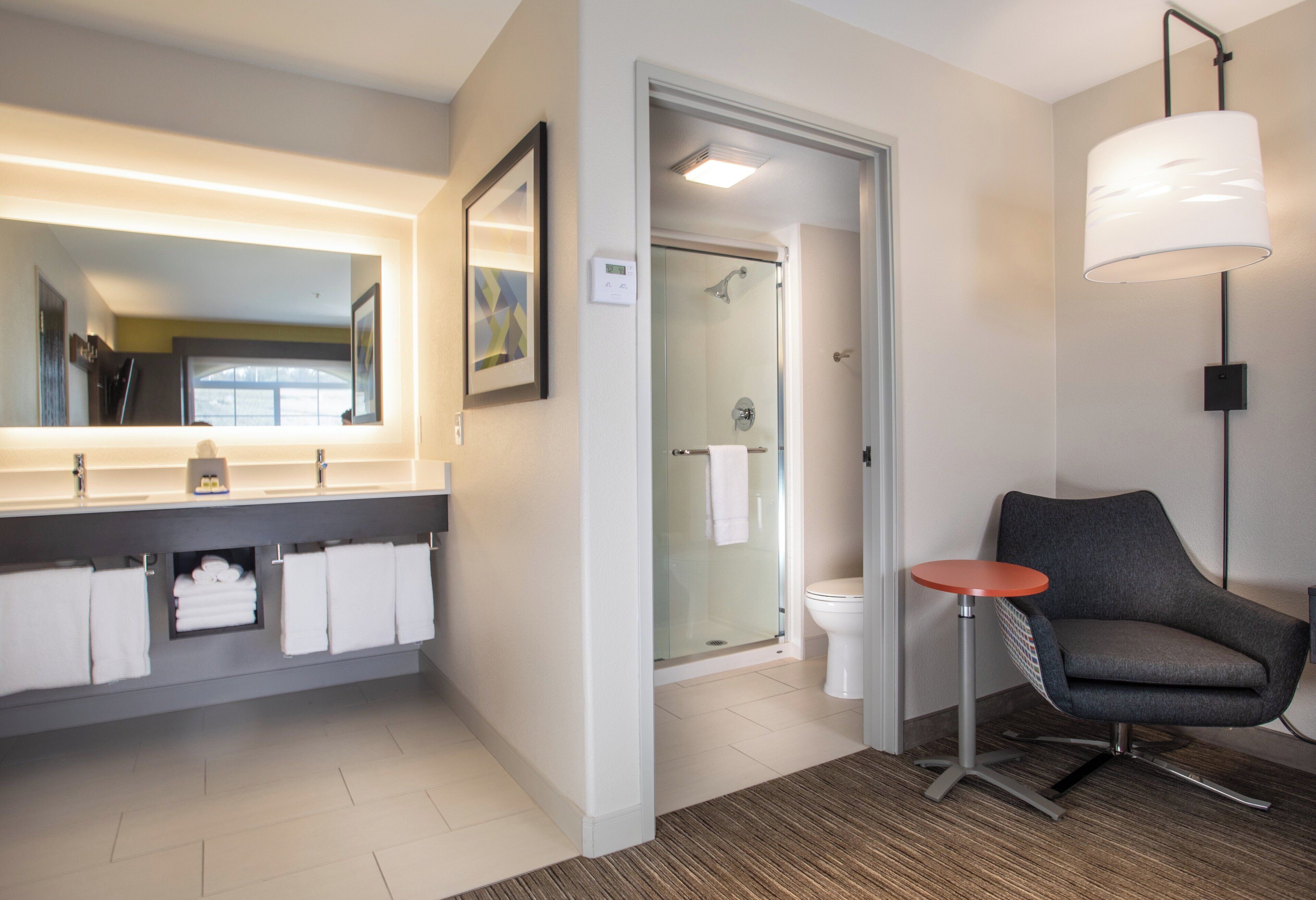 A modern bathroom with a large mirror, a glass shower enclosure, a toilet, and a chair.