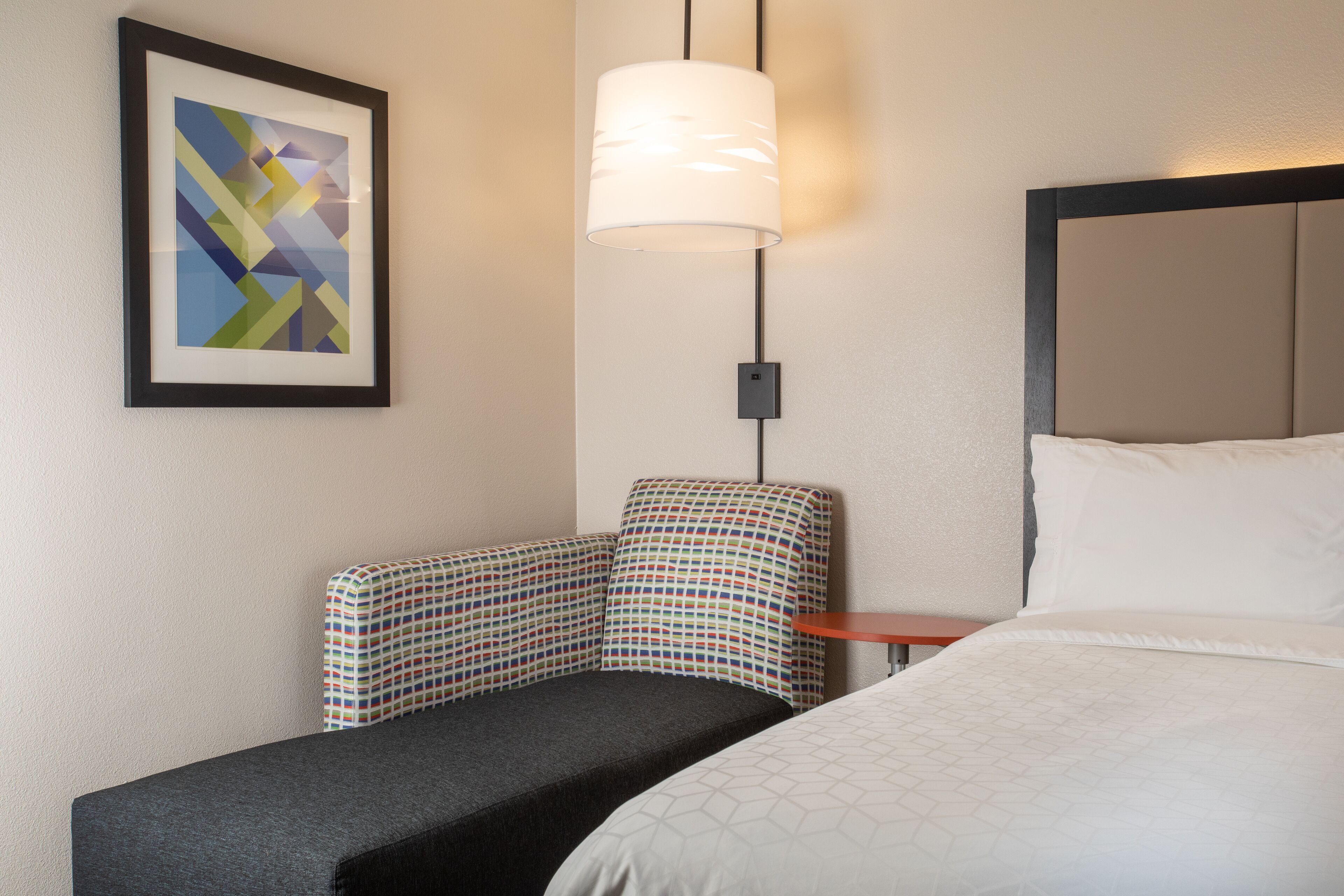 Habitación de hotel con cama, un pequeño sofá, una lámpara de pared y un cuadro abstracto enmarcado.