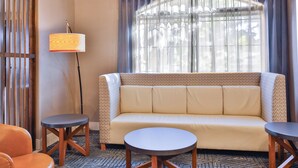 A hotel room with a sofa, two small tables, a lamp, and a window with curtains.