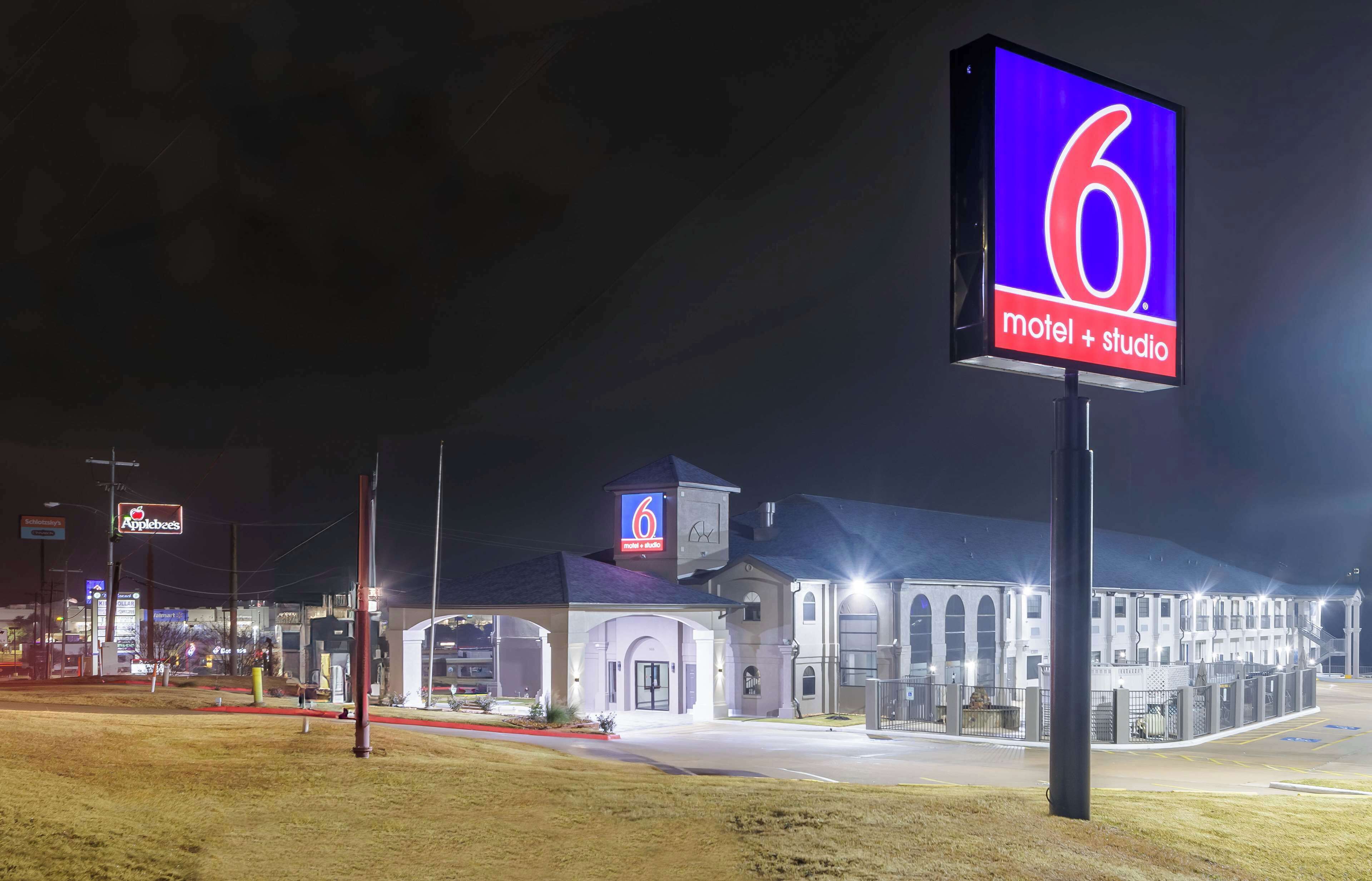Un motel avec un grand panneau indiquant le chiffre « 6 », un vaste parking et un restaurant Applebee’s bien visible à l’arrière-plan.