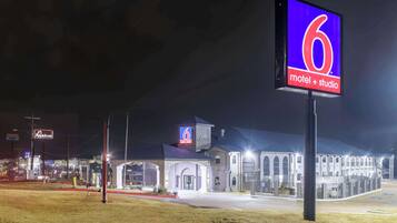 Un motel avec un grand panneau indiquant le chiffre « 6 », un vaste parking et un restaurant Applebee’s bien visible à l’arrière-plan.