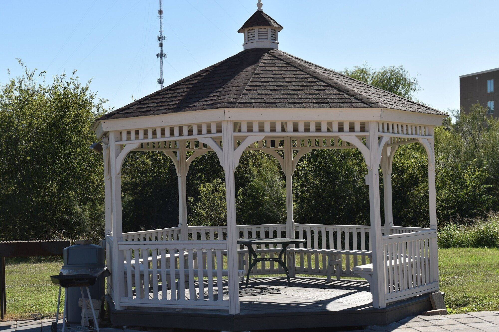 A white gazebo with a peaked roof, surrounded by a white fence, and a table and grill set outside.