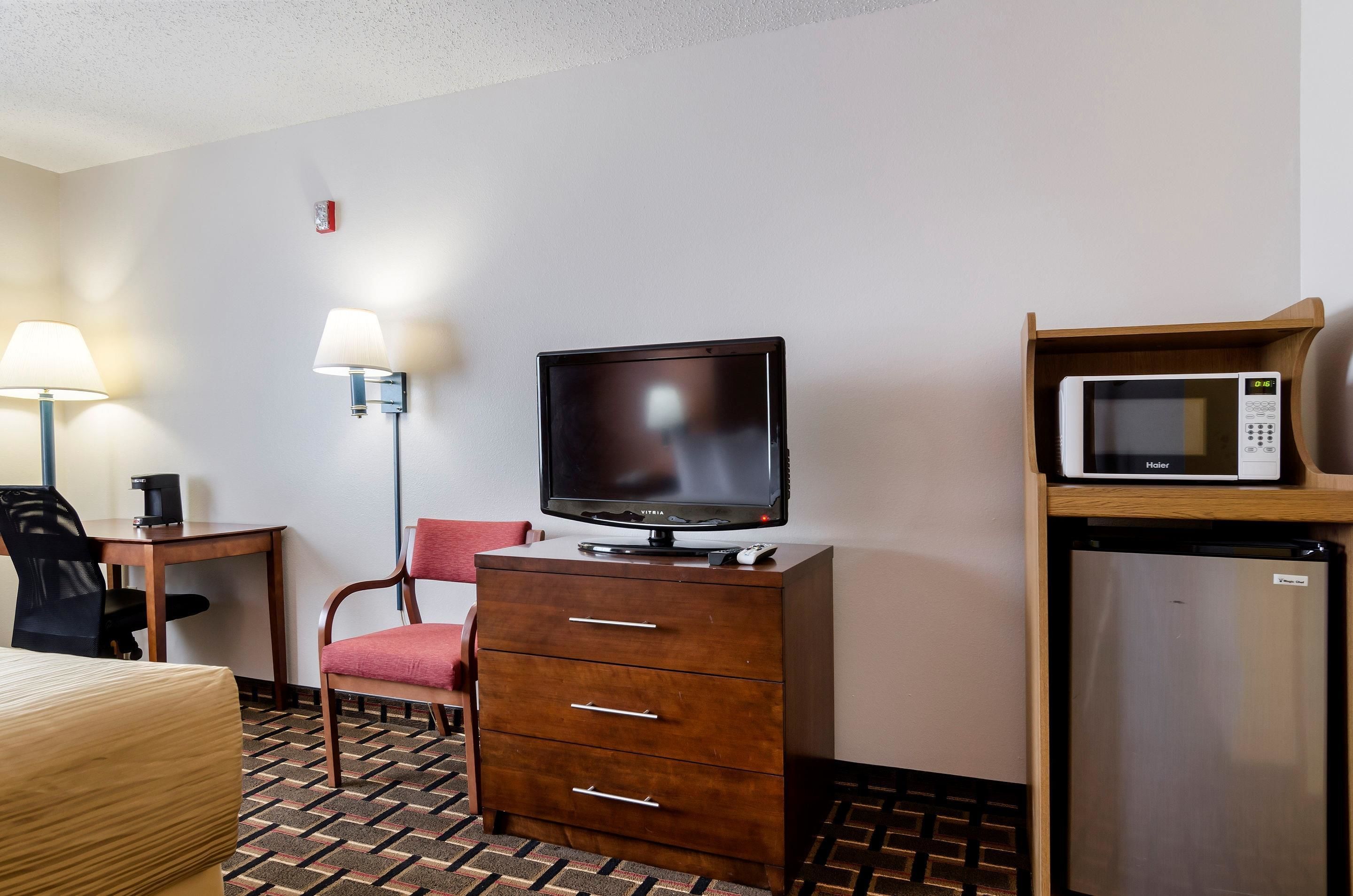 A hotel room with a television on a wooden cabinet, a microwave on a shelf, a desk with a lamp, a chair, and a bed.